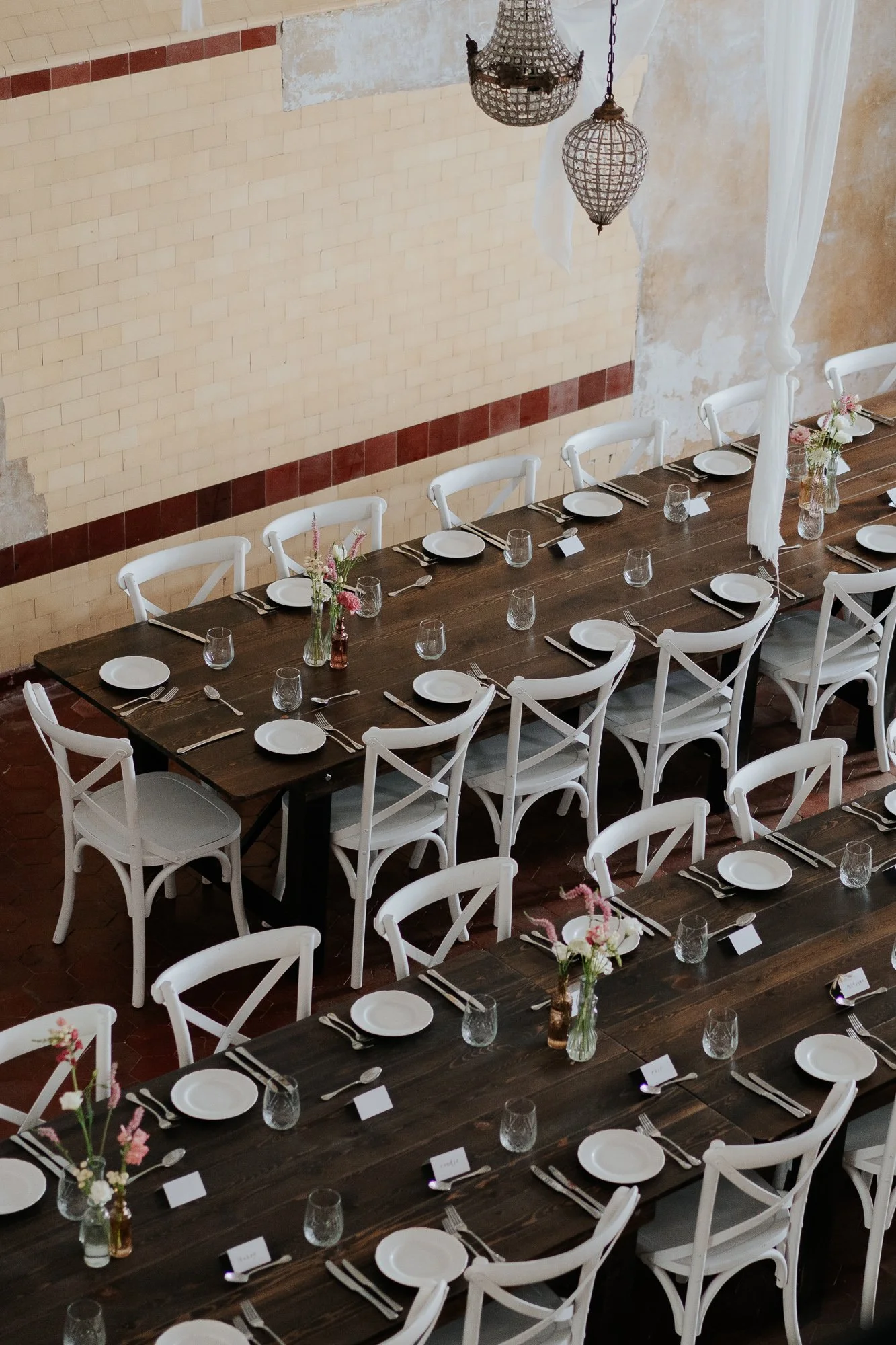 A long dining table set with white plates, glasses, and cutlery, surrounded by white chairs, with floral centerpieces, in a rustic-style room with a brick wall and hanging light fixtures in Amarti at the Heritage Listed Cowwarr Butter Factory.