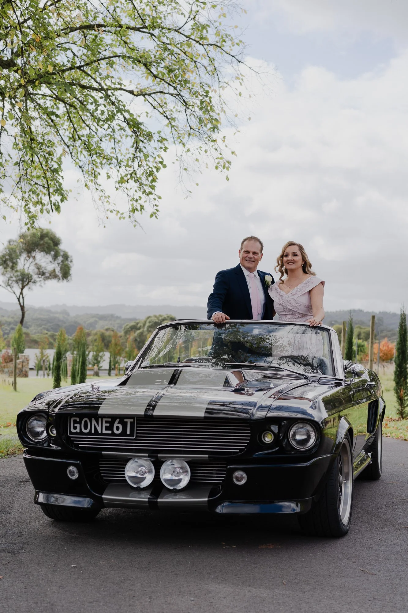 A man and a woman in formal attire standing in a vintage black car with racing stripes outdoors on a cloudy day, with trees and a rural landscape in the background.