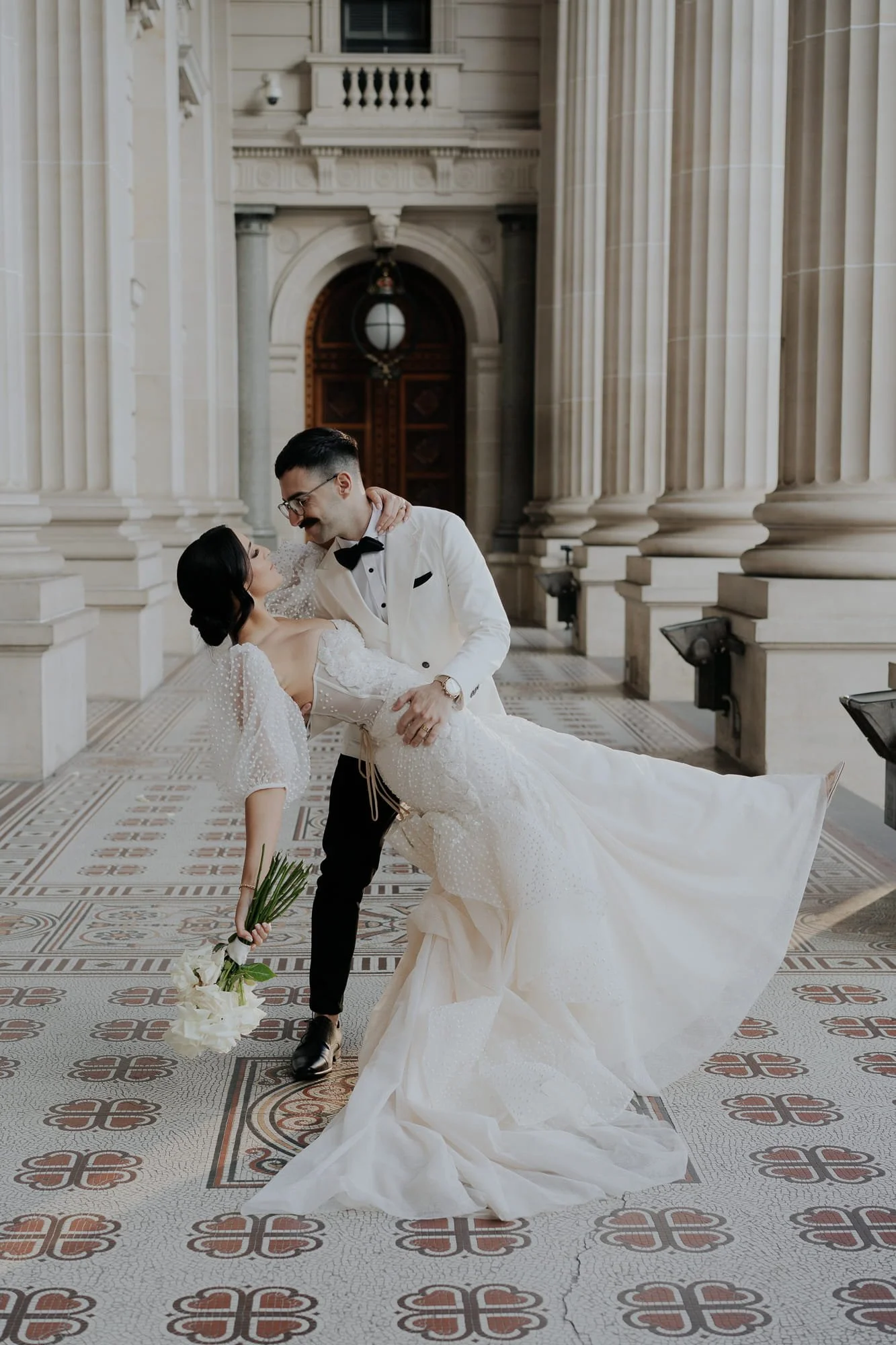 Melbourne Wedding Photography. Parliament House. A bride and groom dancing inside a grand, historic building with tall columns and patterned tile flooring. The groom is wearing a white tuxedo jacket, black bow tie, and black pants, while the bride is