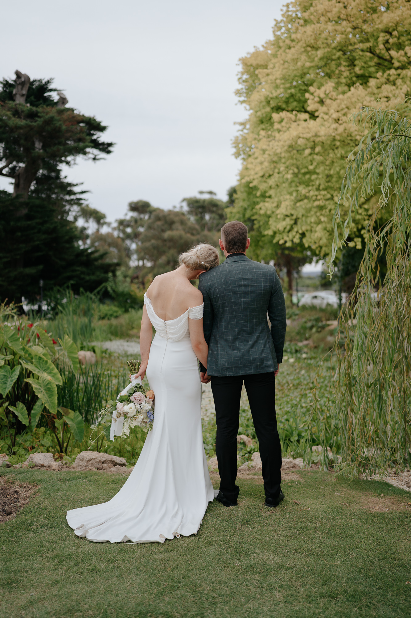 A bride and groom stand together outdoors, facing away from the camera, surrounded by greenery and tall trees, on their wedding day.