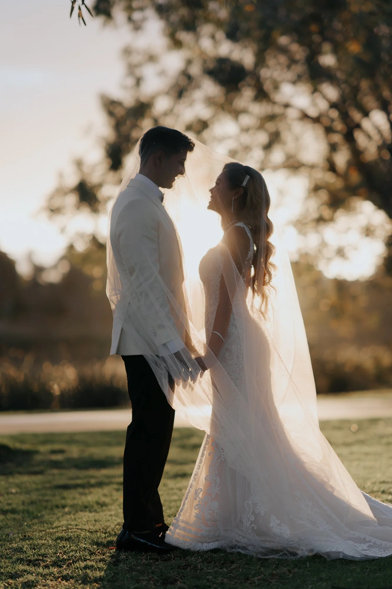 A bride and groom holding hands and facing each other at sunset outdoors, with the bride wearing a lace wedding gown and veil, and the groom in a white jacket and black pants, silhouetted against the sun behind them. Lakeside Events & Conventions