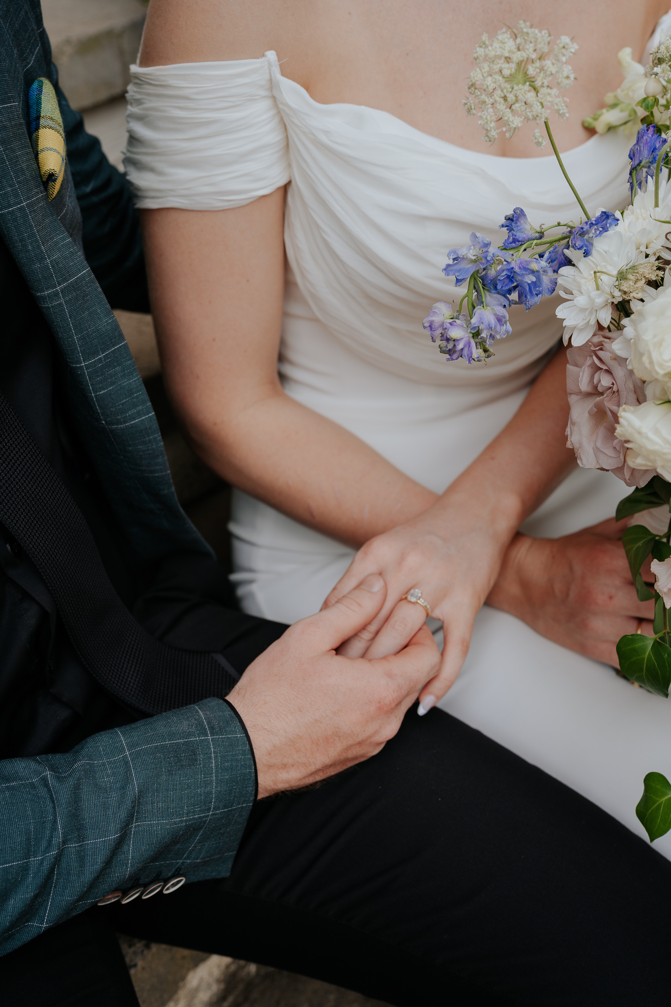 Close-up of a bride and groom holding hands during their wedding, with the groom placing a ring on the bride's finger. The bride is wearing a white off-shoulder wedding dress and holding a bouquet of flowers, including purple, white, and pale pink bl