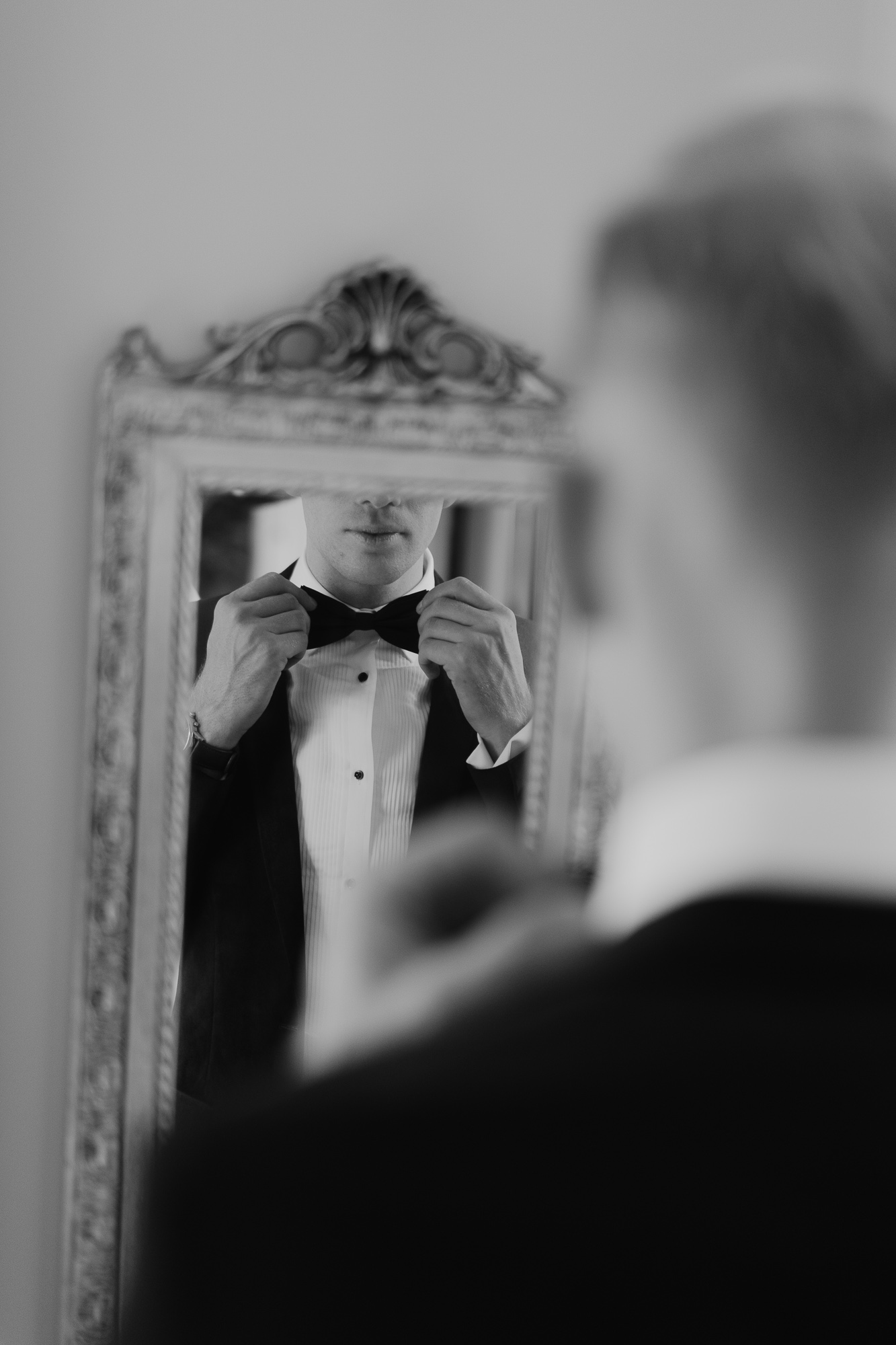 A man dressing in a tuxedo, adjusting his bow tie while looking at his reflection in a decorative wall mirror.