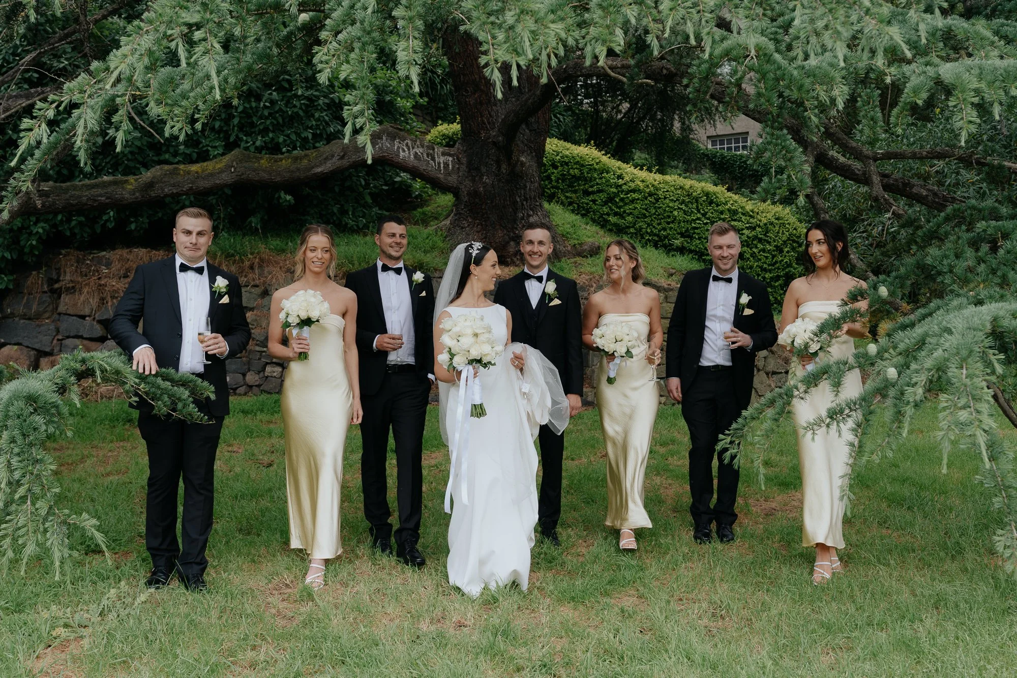 A wedding party of eight people standing outdoors in front of a large tree, with three men in tuxedos and five women, four in butter-colored dresses and one in a white wedding gown, holding bouquets and glasses. Leonda by the Yarra