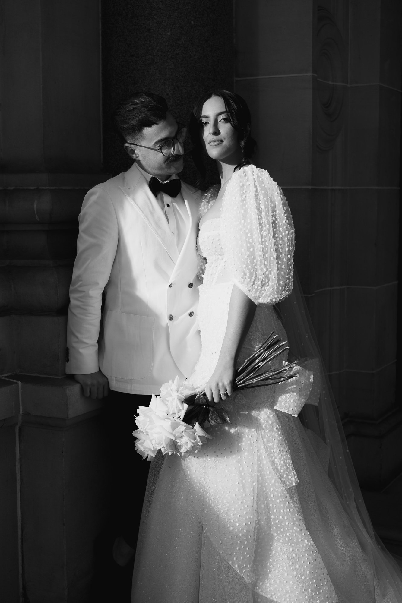 A black-and-white photo of a couple dressed in wedding attire, with the man in a tuxedo and the woman in a wedding gown holding a bouquet of flowers. They are standing against a stone wall at Parliament House, Victoria.