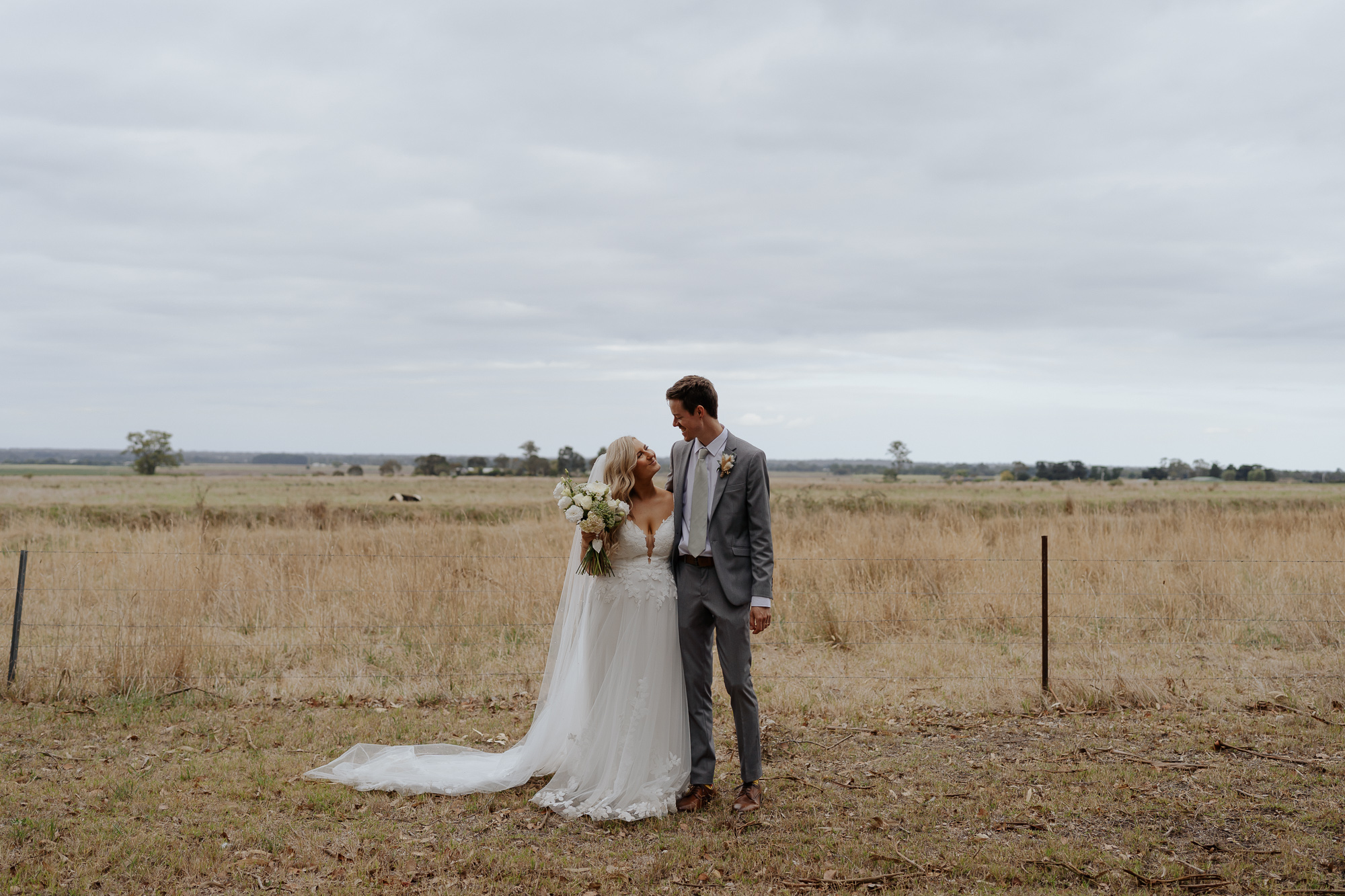 A bride and groom standing together in a field, looking at each other, with the bride holding a bouquet of white flowers, overcast sky in the background. The Vines on Avon