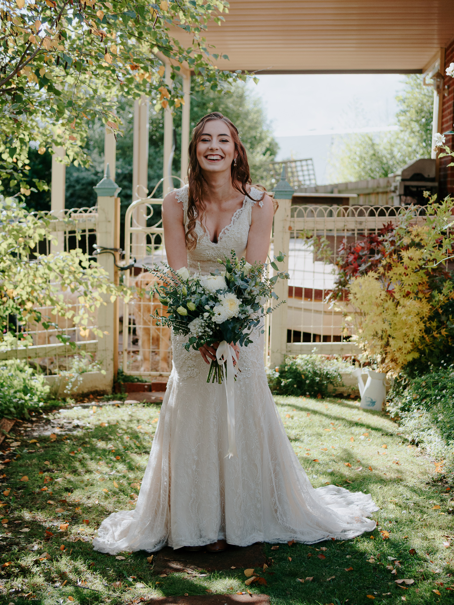 A bride in a white lace wedding dress holding a bouquet of white flowers and greenery, standing outdoors in a garden with trees and a wooden fence.