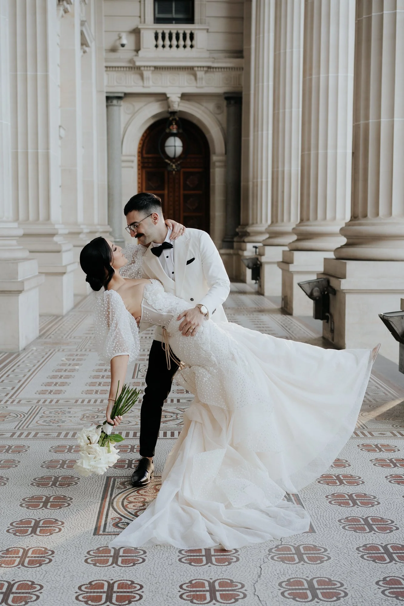 A bride and groom dancing in a grand, marble hall with large columns and ornate floor tiles. The bride is dressed in a white wedding gown and holding a bouquet, while the groom is in a white tuxedo with a black bow tie.