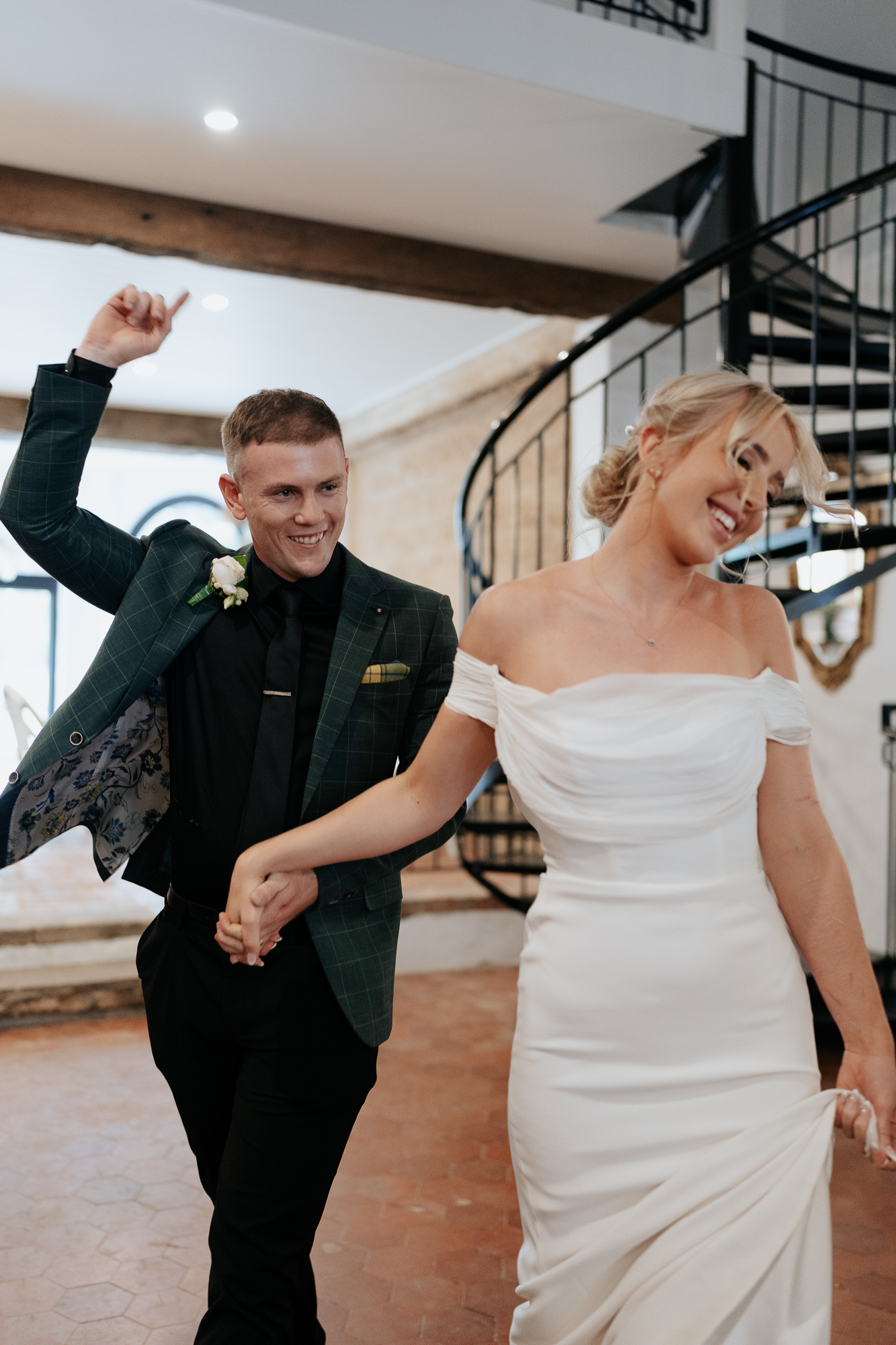 A joyful couple holding hands and dancing indoors, with the man raising one arm up, on a wedding day.