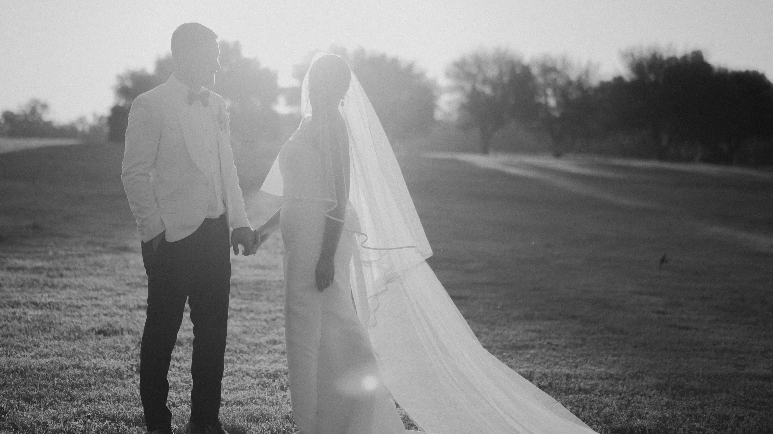 A couple holding hands on a grassy field with trees in the background, dressed in wedding attire, with the woman wearing a veil and the man wearing a suit and bow tie, during sunset.