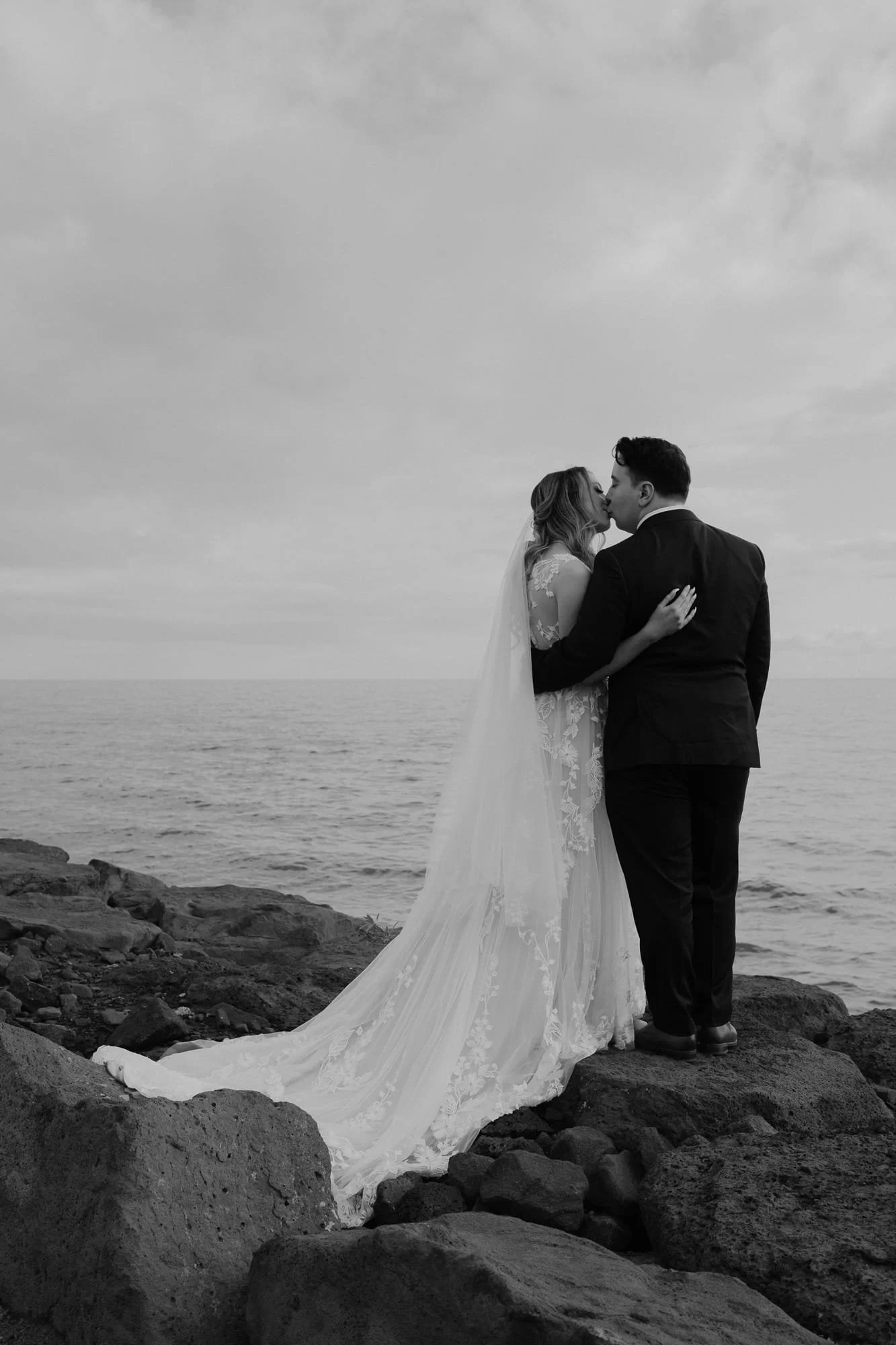 Melbourne Wedding Photography. Black and white photograph of a bride and groom sharing a kiss on a rocky shoreline behind Sandringham Yacht Club, with the ocean and cloudy sky in the background.