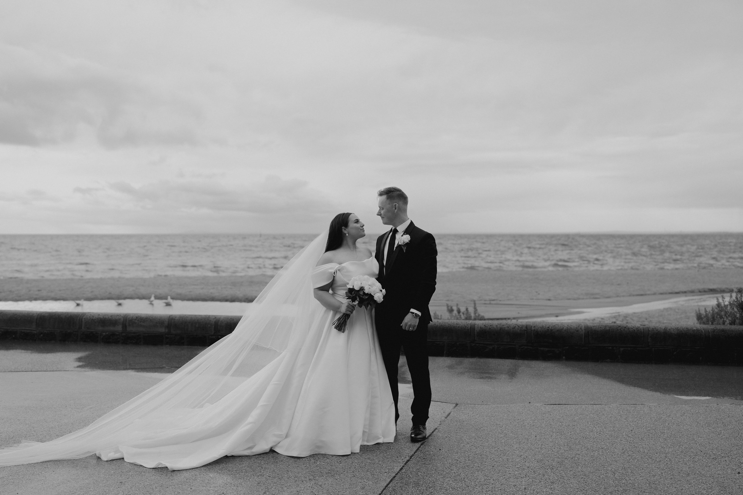 Black and white photo of a bride and groom standing on a promenade by the ocean, the bride holding a bouquet of flowers, wedding gown with a long train and veil, groom in a suit with boutonniere, both gazing at each other.