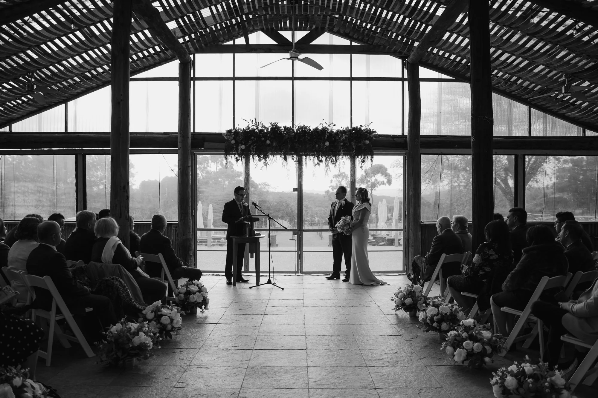 Black and white photo of a wedding ceremony inside a rustic wooden venue with large windows, showing a bride and groom standing before an officiant, guests seated on either side, and floral arrangements along the aisle.