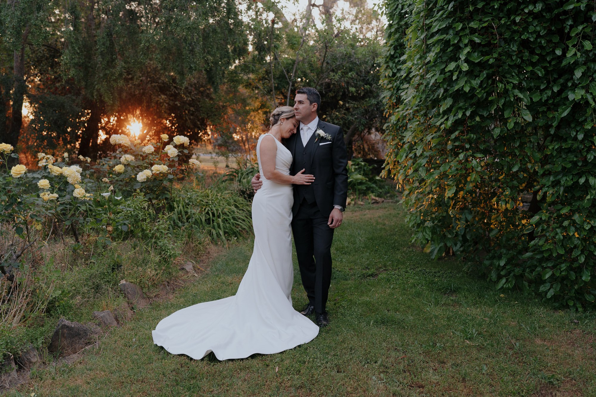 A bride and groom embrace in a garden at sunset, with the bride wearing a white gown and the groom in a black suit. Wedding held at Buller Wines, Rutherglen.