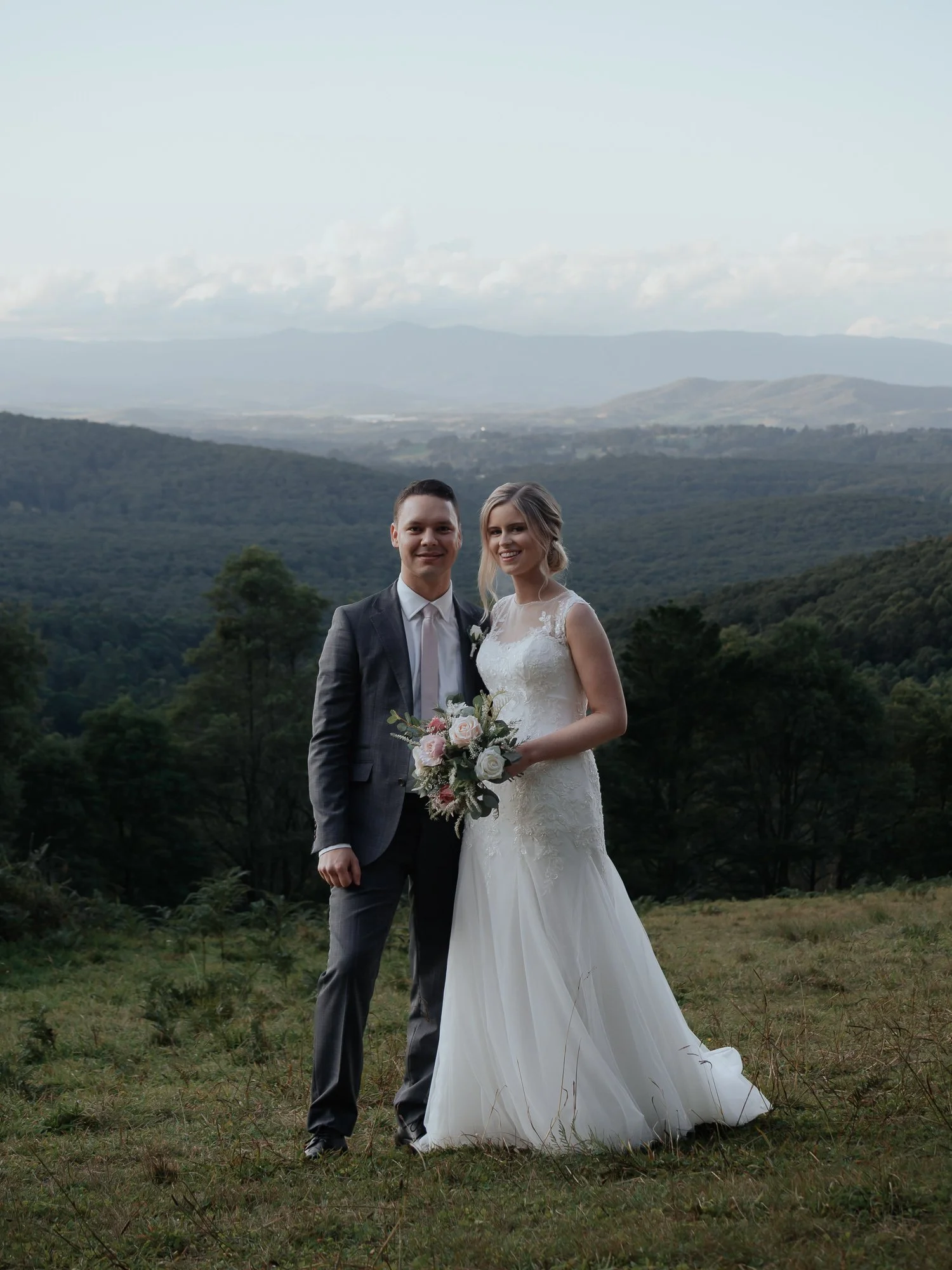 A bride and groom standing outdoors on a grassy field with a mountain and forested landscape in the background, dressed in wedding attire.