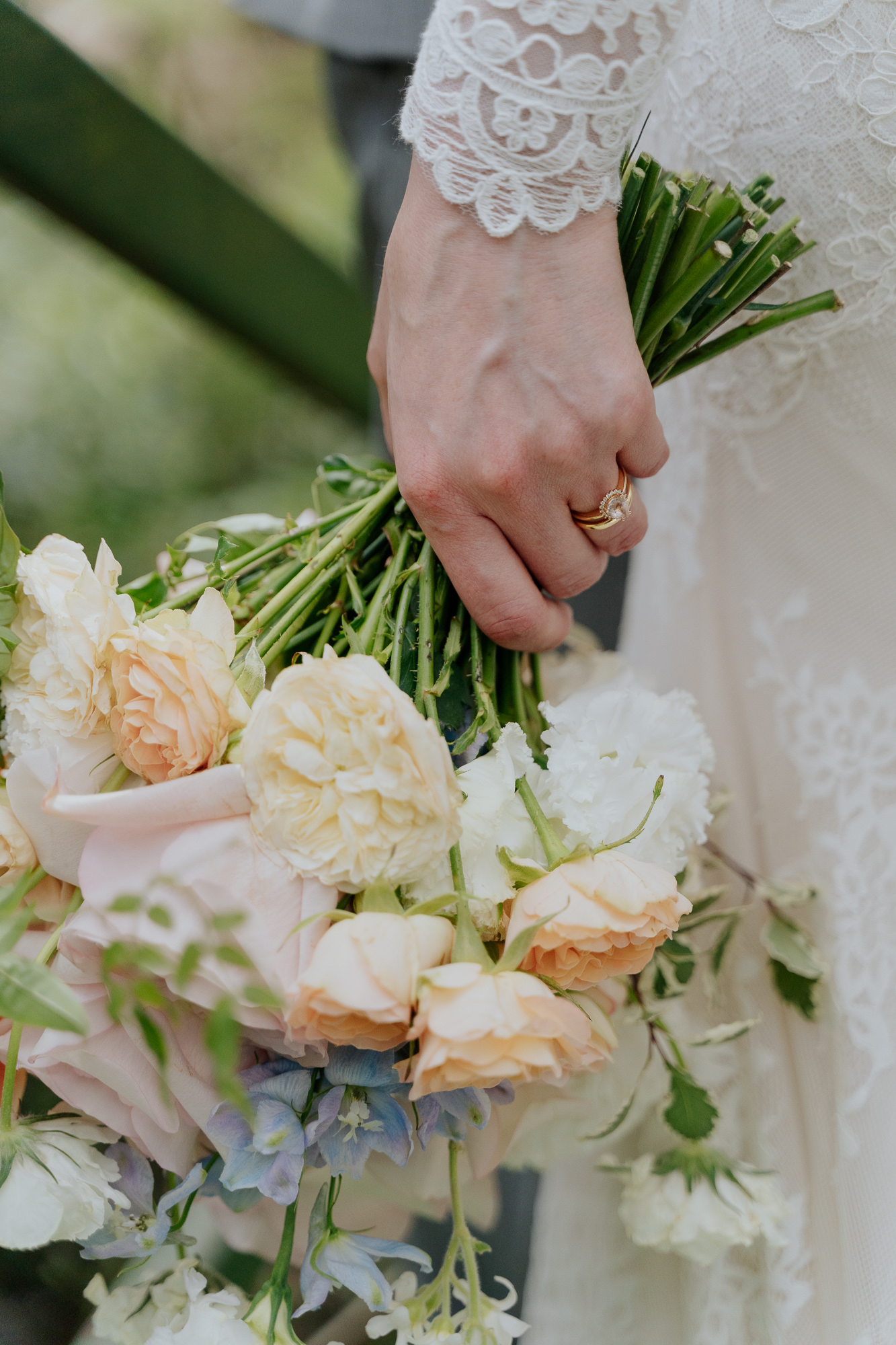 Close-up of a woman's hand holding a bouquet of soft-colored flowers, wearing a gold ring, and dressed in white lace fabric.