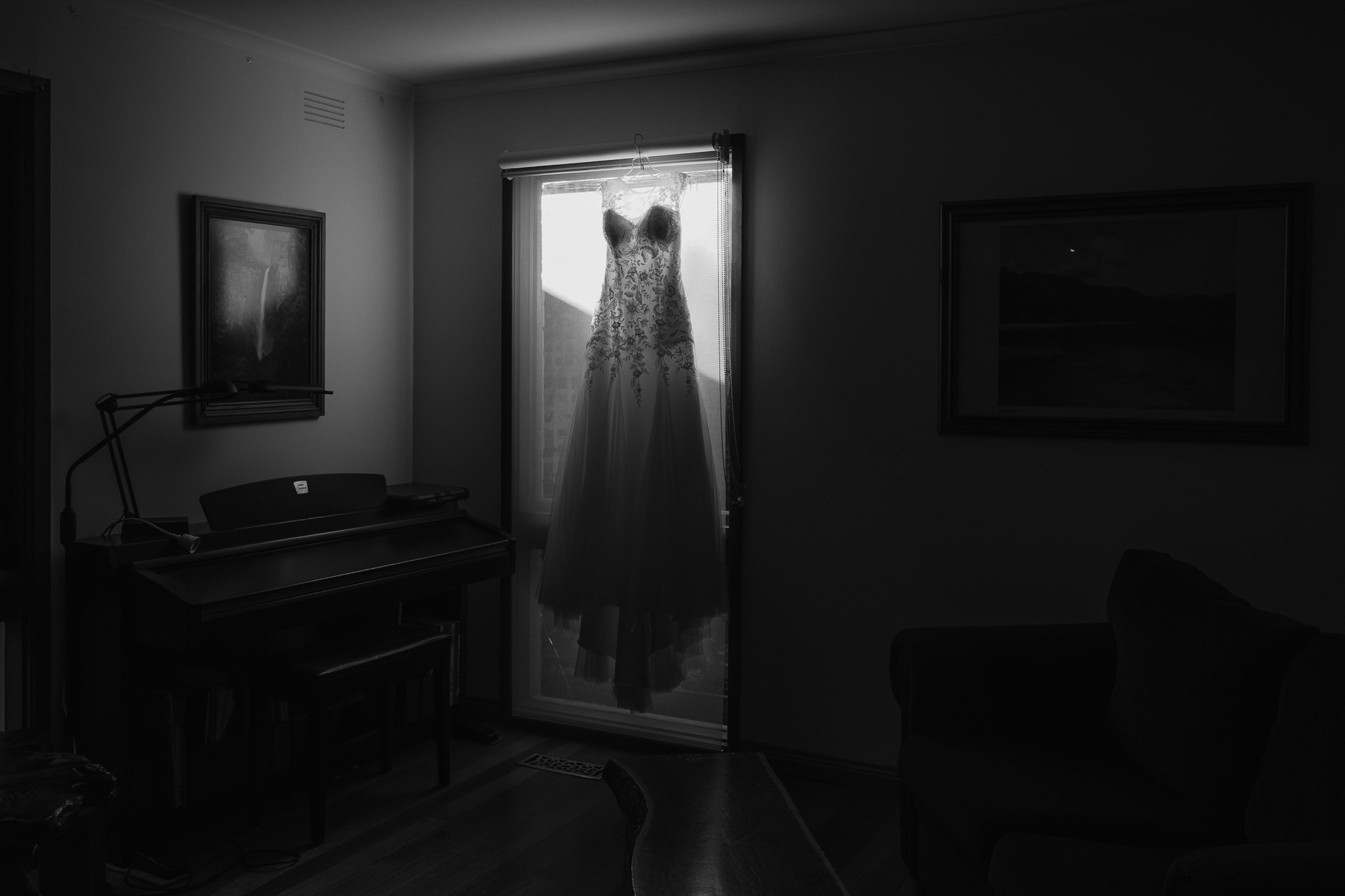 A wedding dress hangs in front of a window in a dimly lit room, with a piano, framed artwork, and a sofa visible.