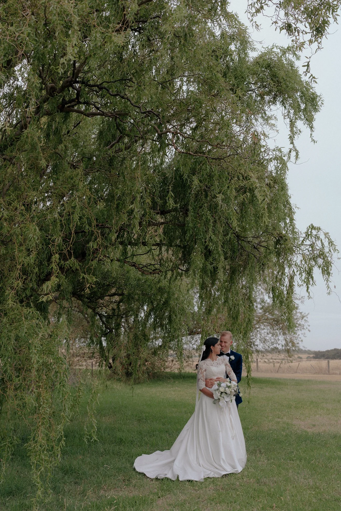 A bride and groom standing together under a large leafy tree in an open field at Witchmount Estate, with the bride holding a bouquet of white flowers.