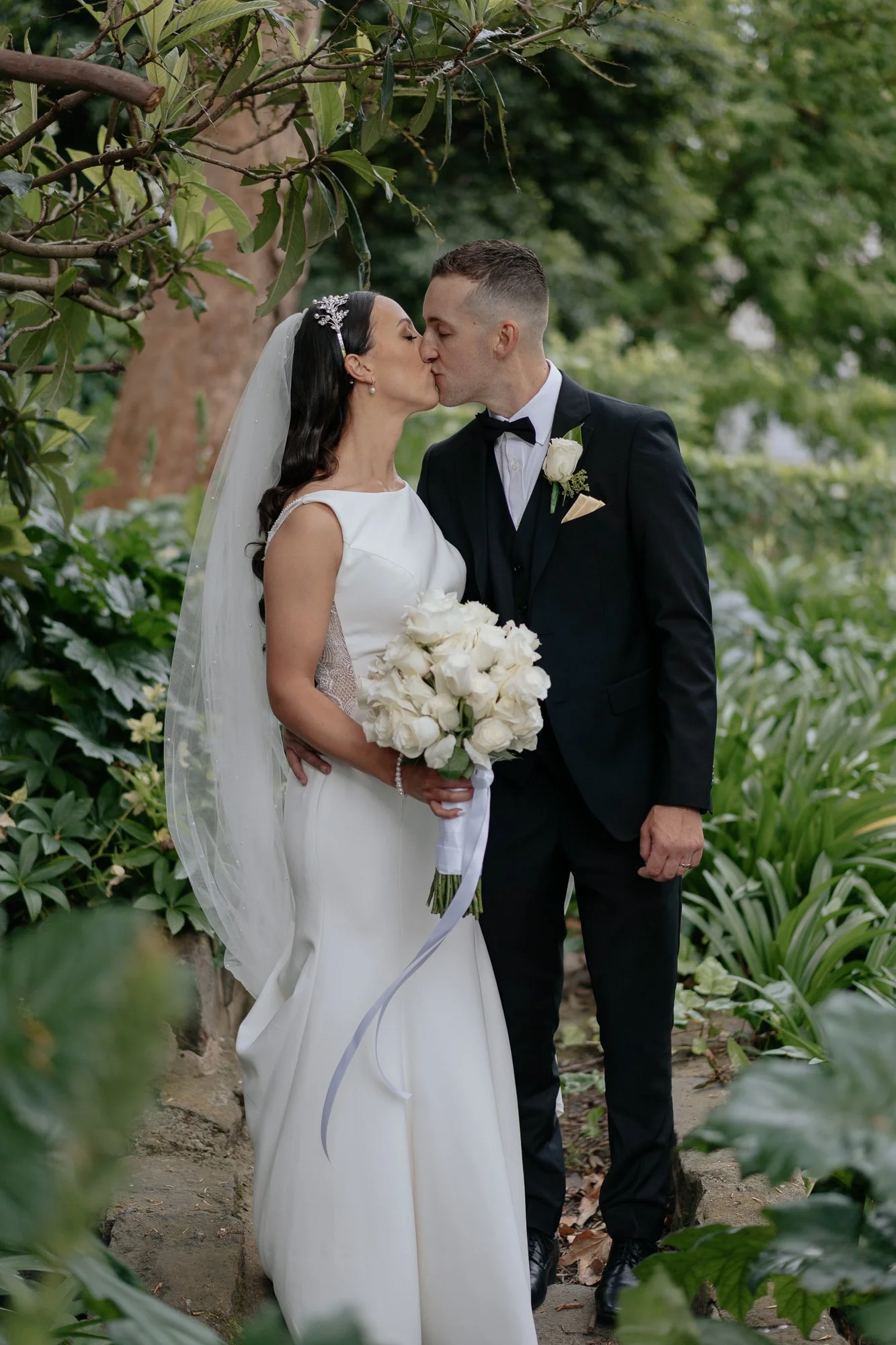 A bride and groom kissing at Leonda by the Yarra, surrounded by greenery, with the bride holding a bouquet of white roses.
