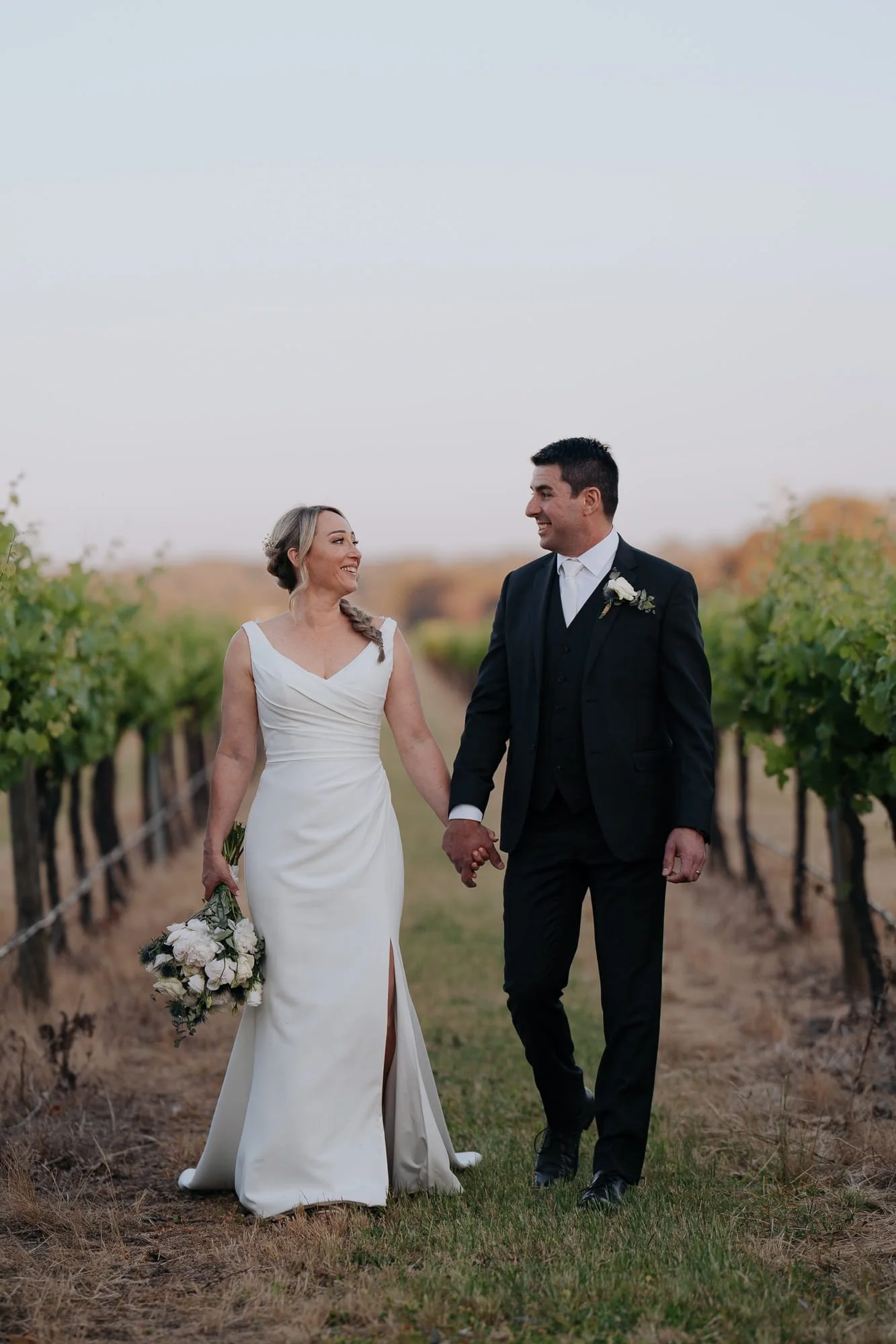 Melbourne Wedding Photography. A bride and groom walking hand in hand through a vineyard at Buller Wines Rutherglen, smiling at each other. The bride is holding a bouquet of flowers, and the groom is dressed in a black suit. It is sunset.