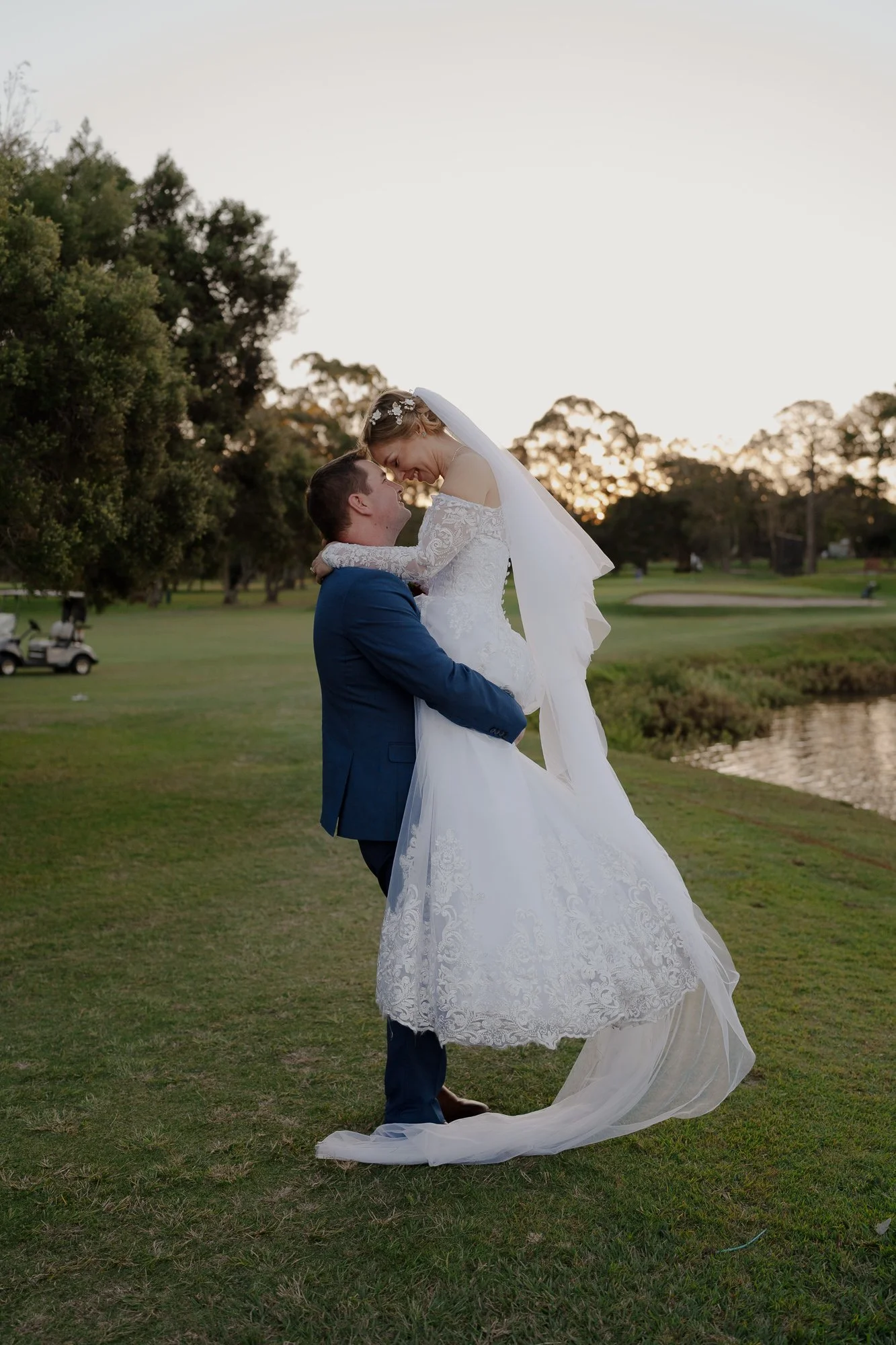 A bride and groom celebrating on Wynnum golf course during sunset, with the groom lifting the bride, who is wearing a white wedding dress and veil, and the groom wearing a blue suit.