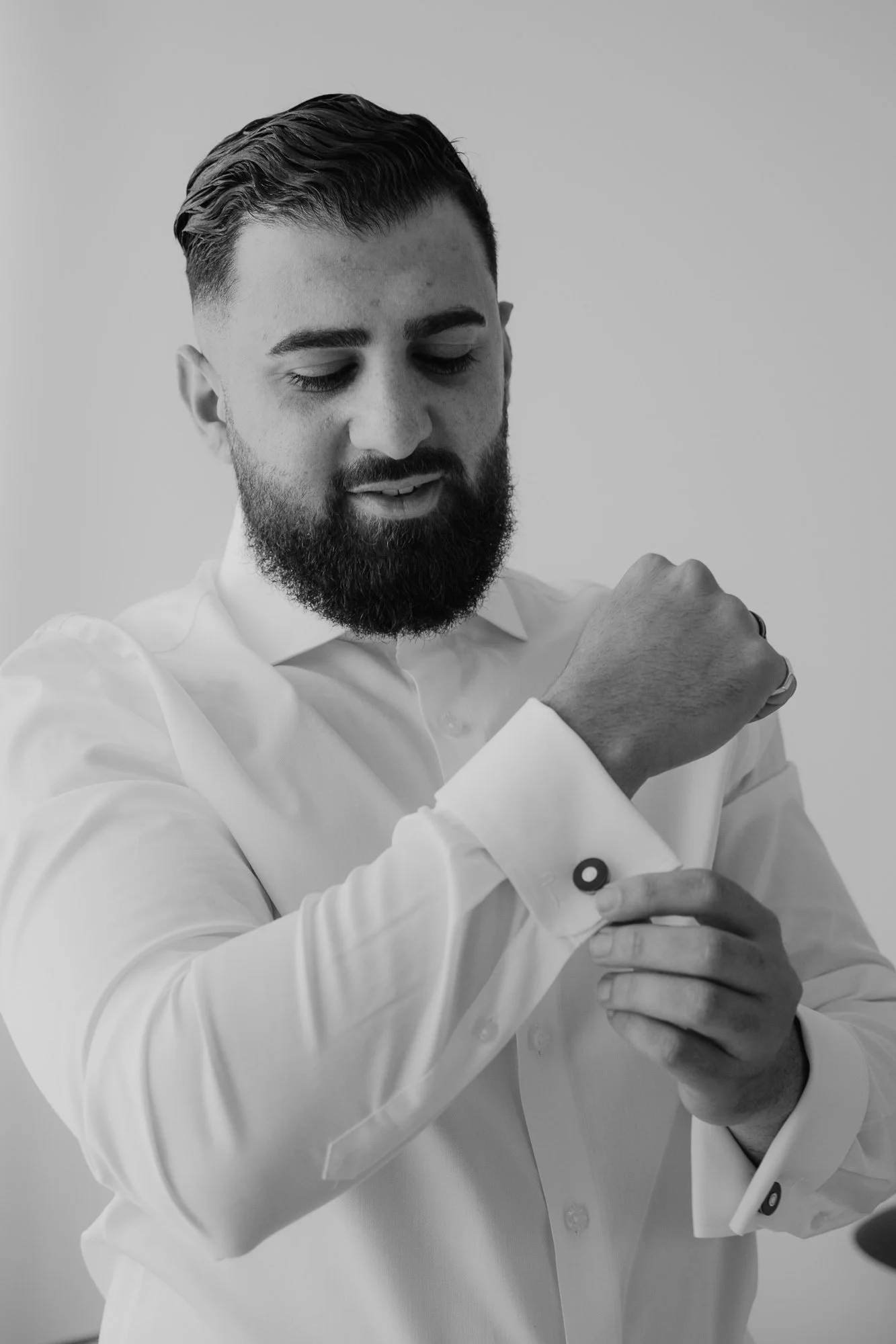 A man in a white dress shirt adjusting his cufflinks, looking down at his wrist in a black and white photo.