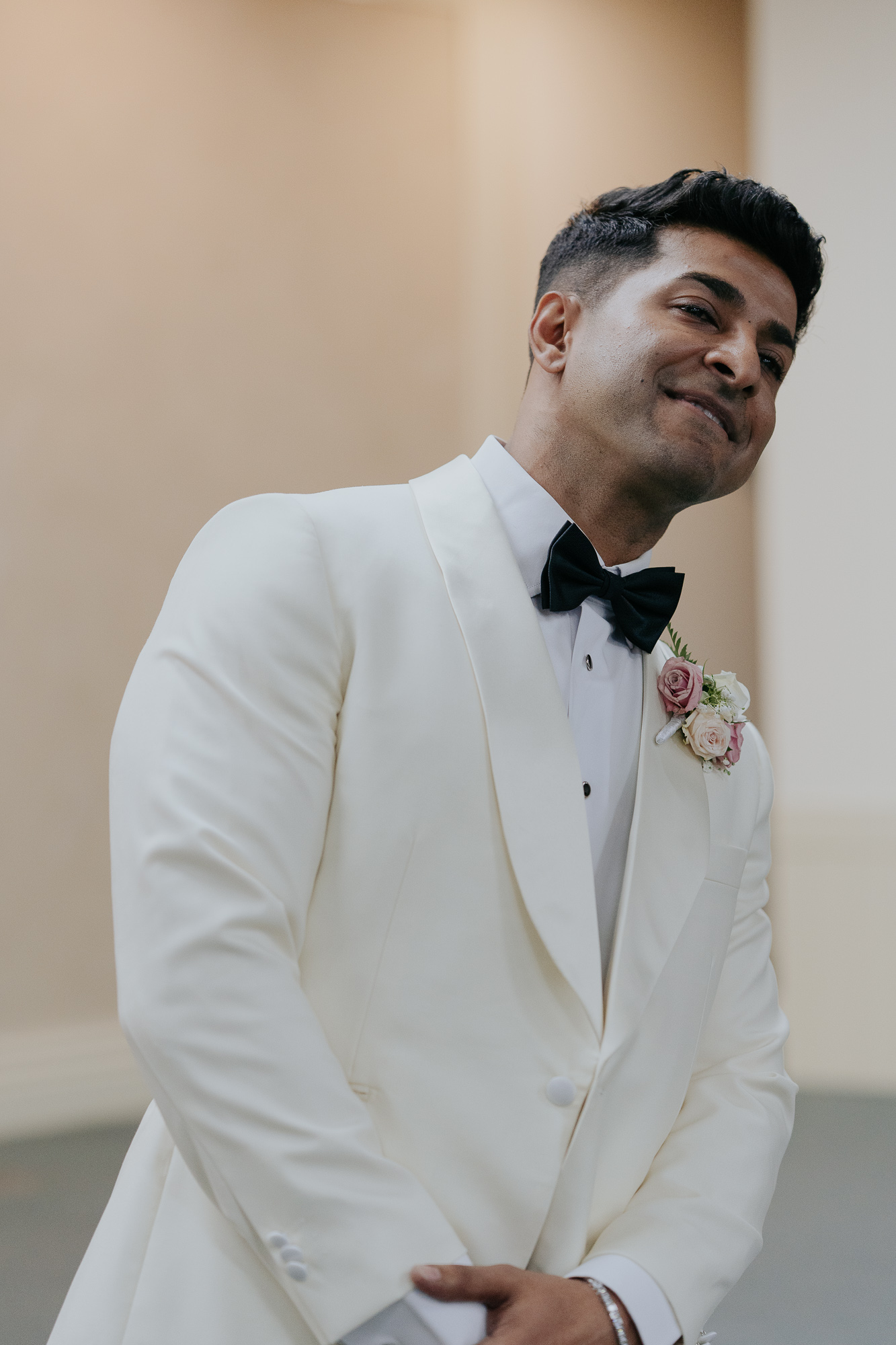 A man dressed in a white tuxedo with a black bow tie, wearing a pink and white boutonniere, smiling softly at a wedding, first look of bride.