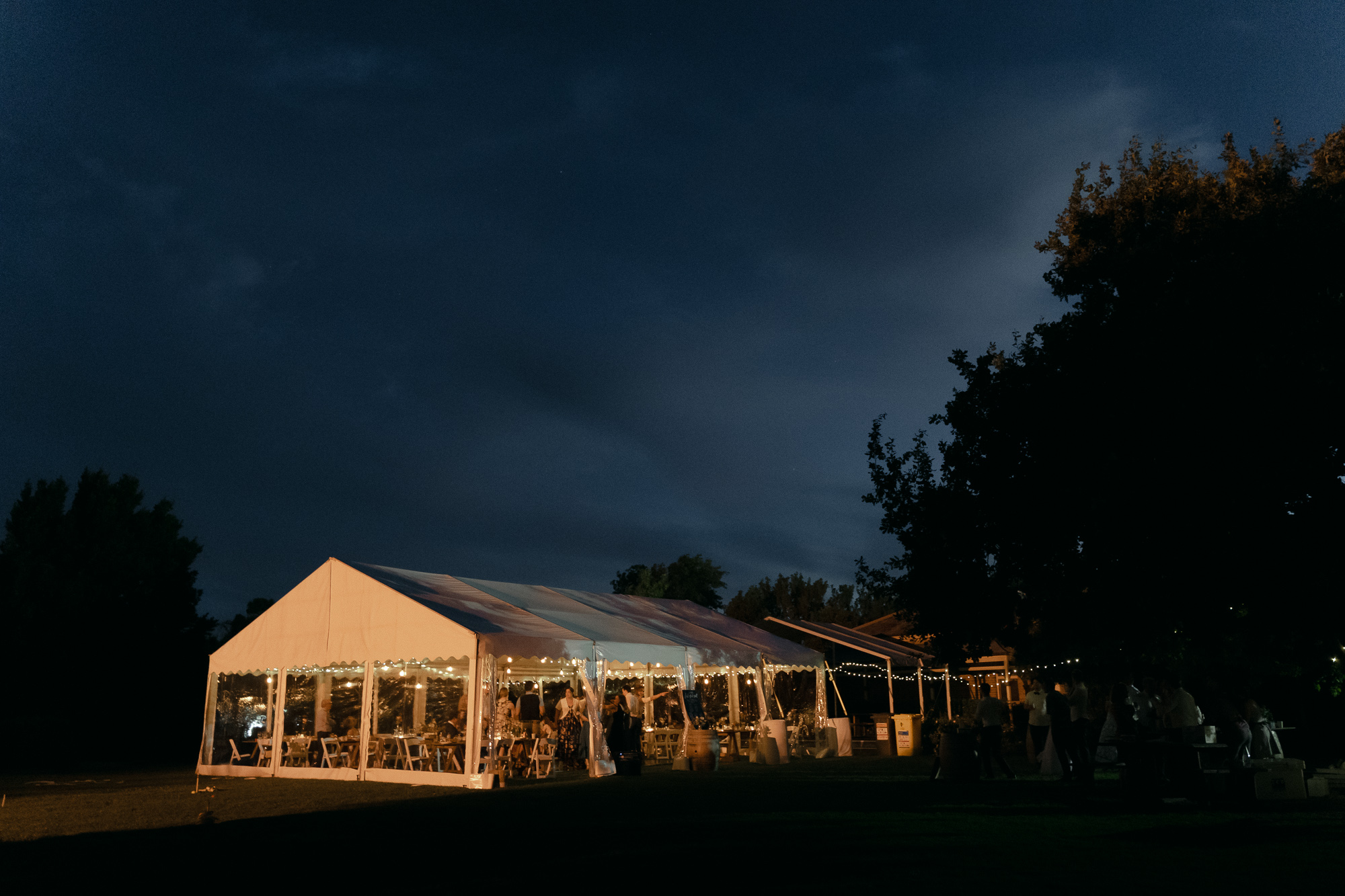 Outdoor evening gathering under a large illuminated tent with people socializing and dining, surrounded by trees and string lights on a dark night sky.