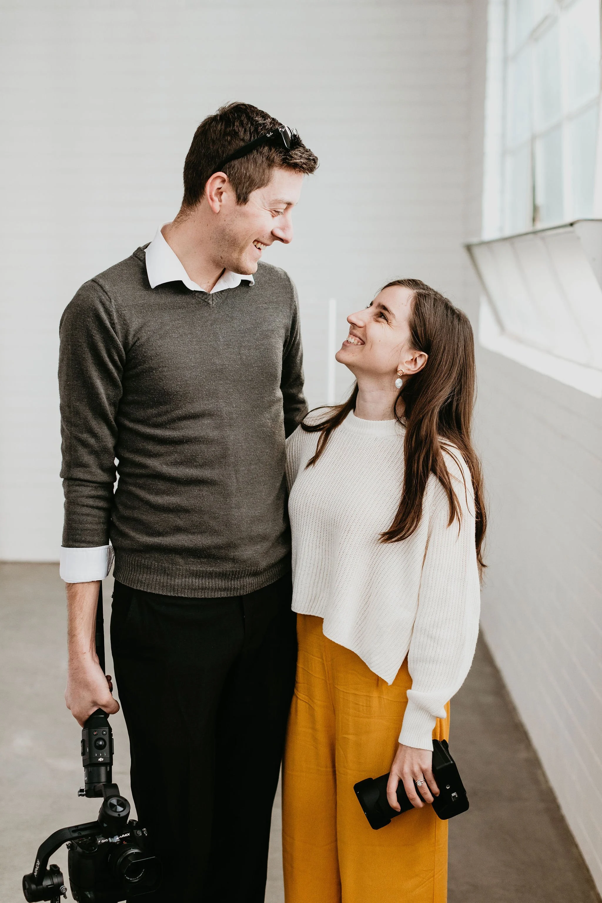 A man and woman smiling and looking at each other indoors, woman holding a camera, man holding a camera lens, bright natural light from a window.