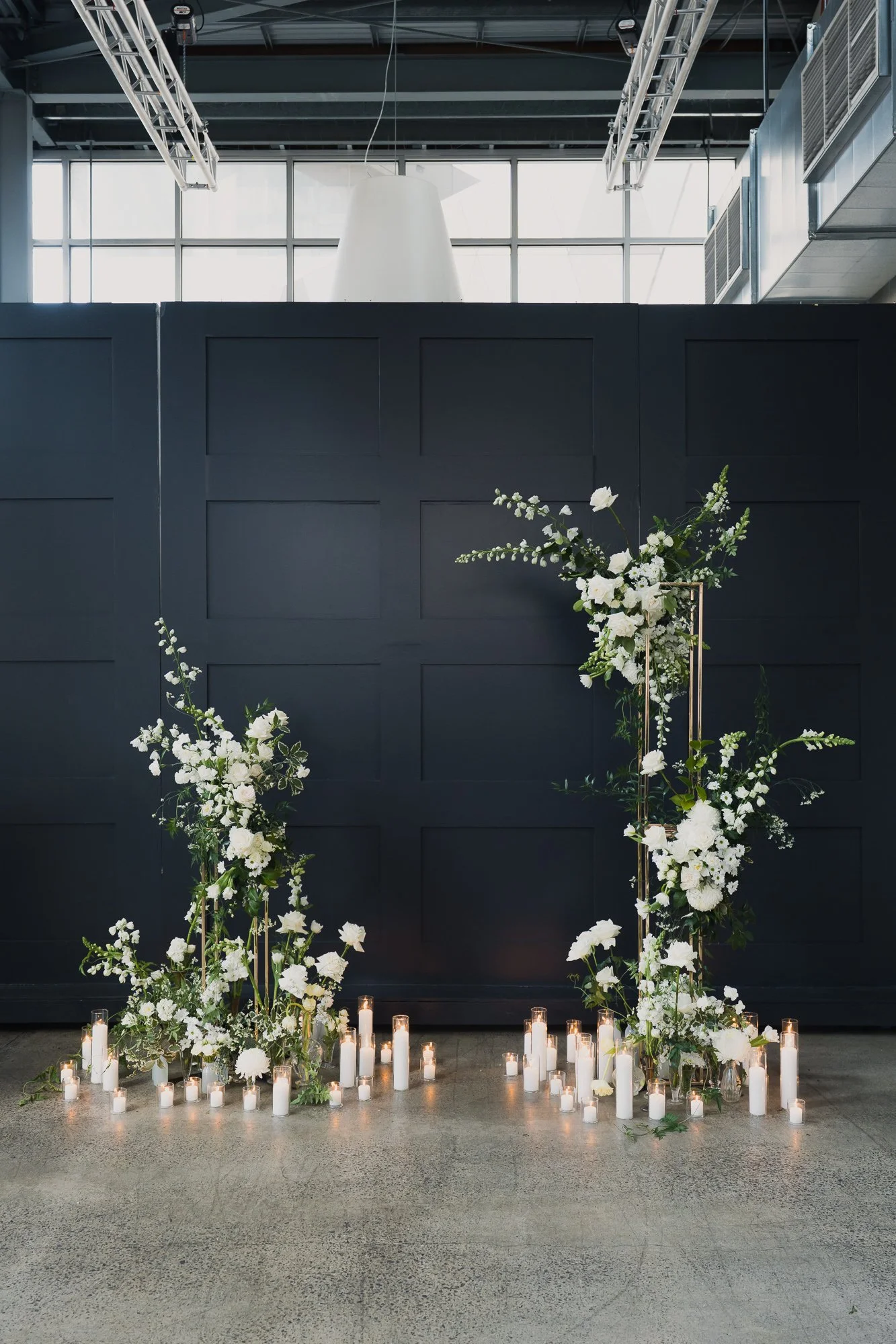 Indoor event space with a dark wall backdrop decorated with white floral arrangements on gold stands and numerous white pillar candles of varying heights on the floor. Alto Event Space, a Melbourne function venue with city views.