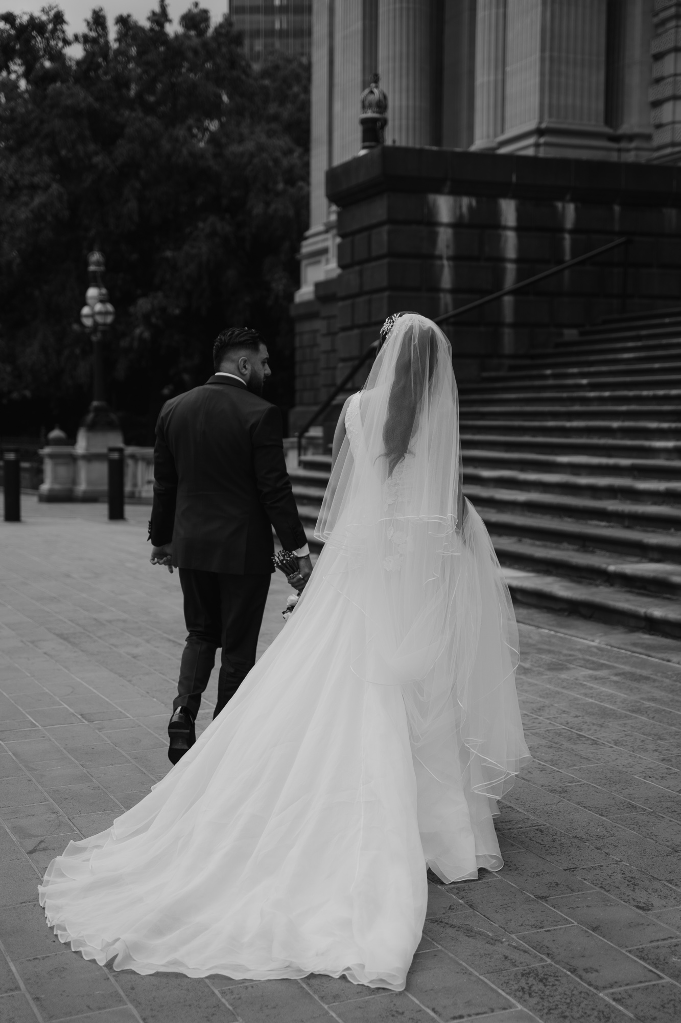 A bride and groom walking hand in hand down stairs outside a large building, with the bride wearing a long wedding gown and veil, and the groom in a dark suit. Parliament House Melbourne Wedding Photography.