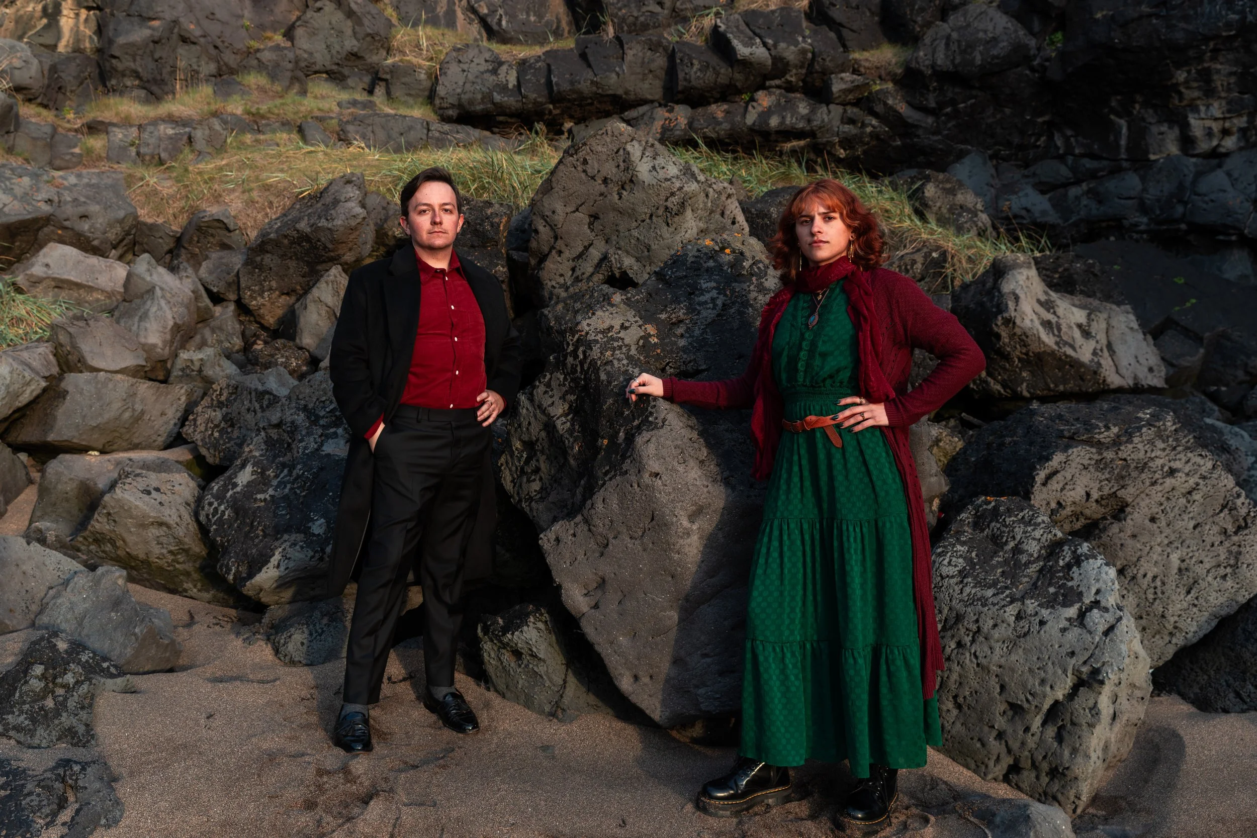 A man and woman standing on a sandy beach with large rocks in the background. The man is dressed in a black coat, red shirt, and black pants. The woman is wearing a green dress, red cardigan, and black boots, with one hand on her hip and the other re