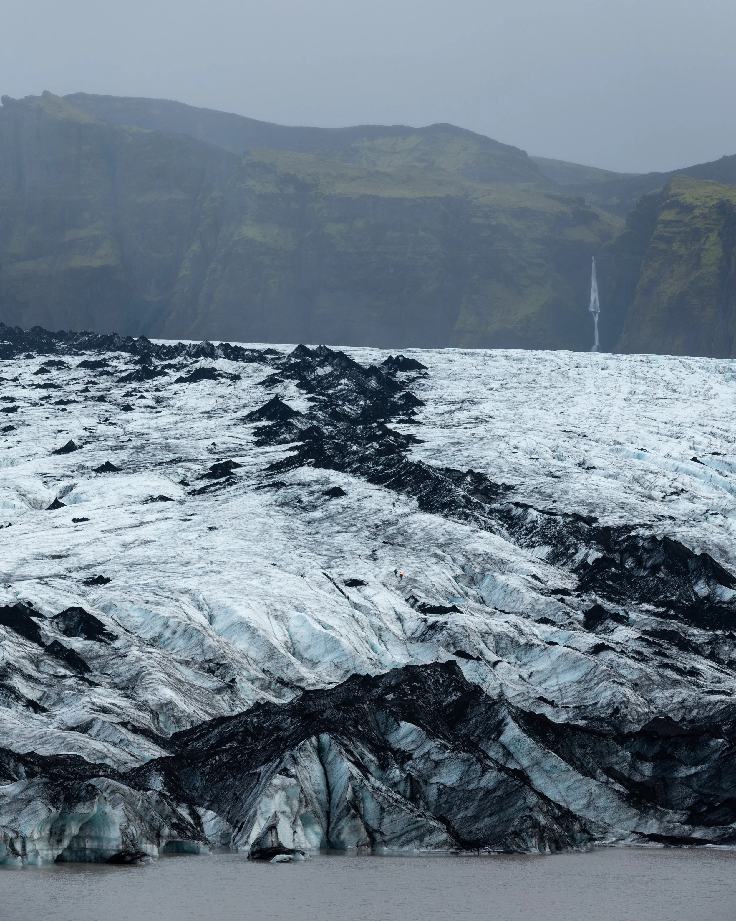 A glacier with black and white ice in a remote mountain landscape, with cliffs and a waterfall in the background.