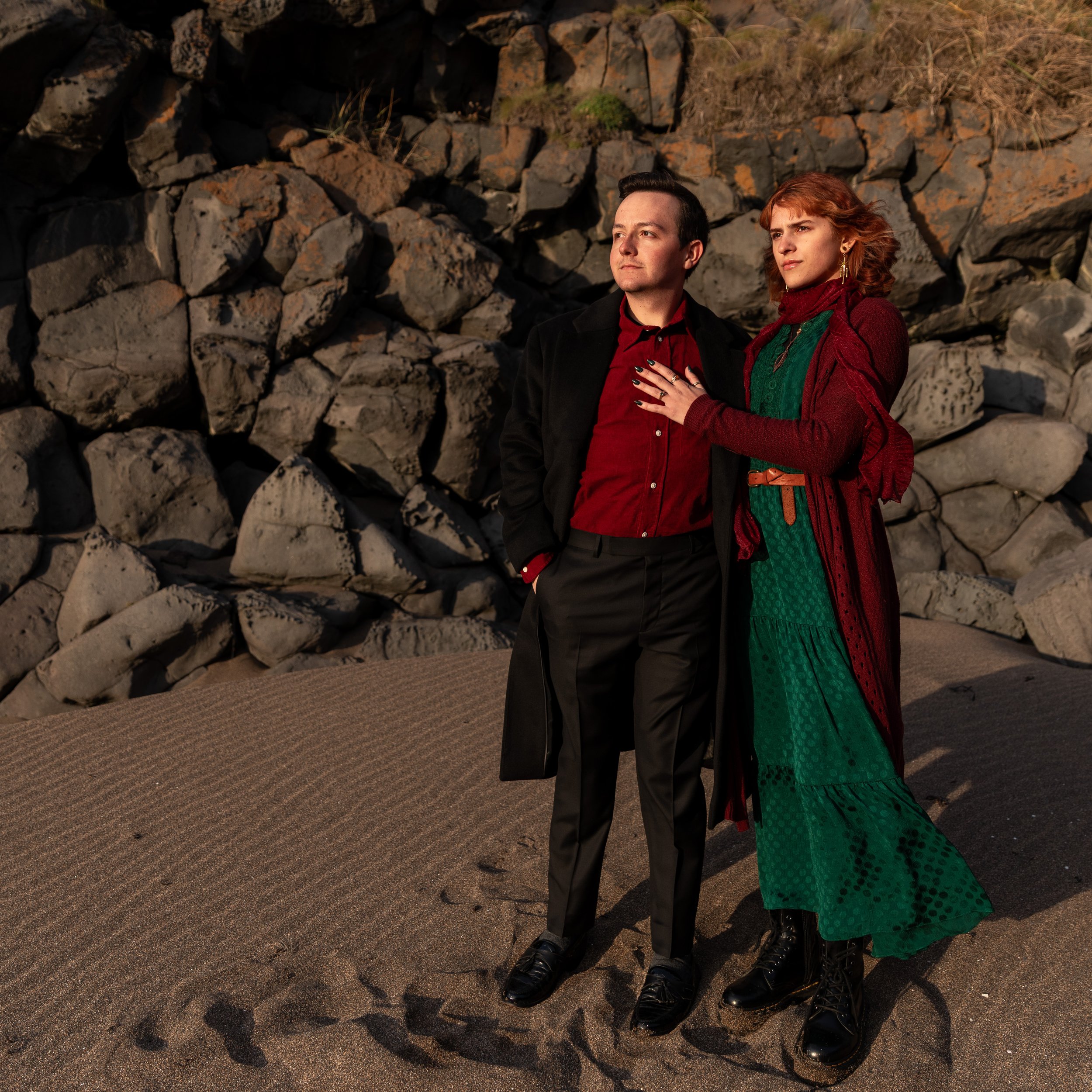 A man and woman standing on sandy ground near rocks, dressed in vintage clothing, looking pensively into the distance.