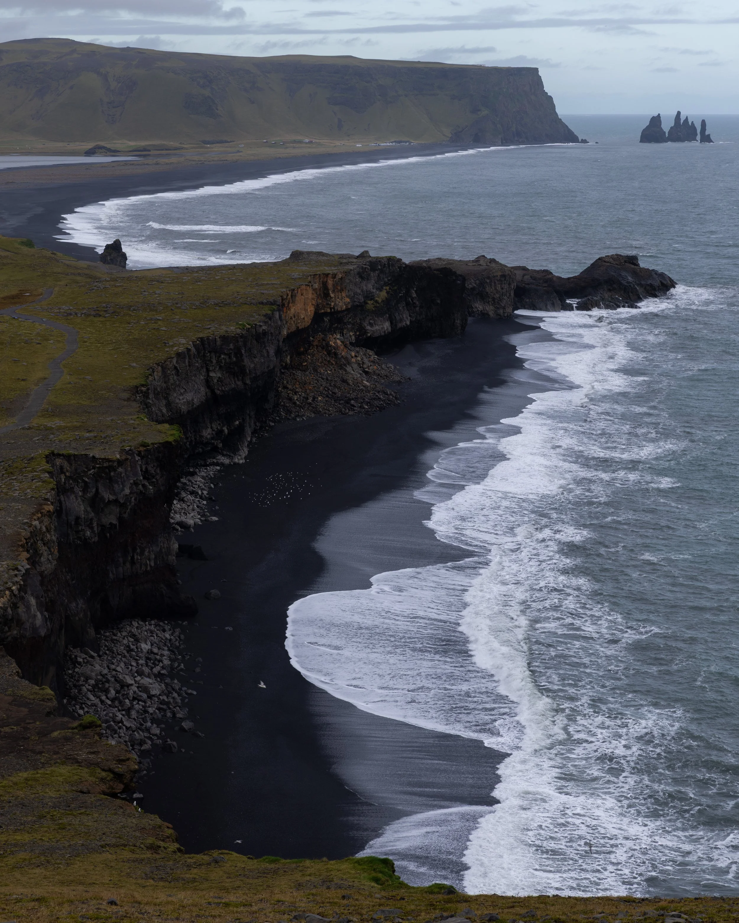 Cliffs overlooking a black sand beach on a cloudy day, with waves crashing and distant rock formations in the ocean.