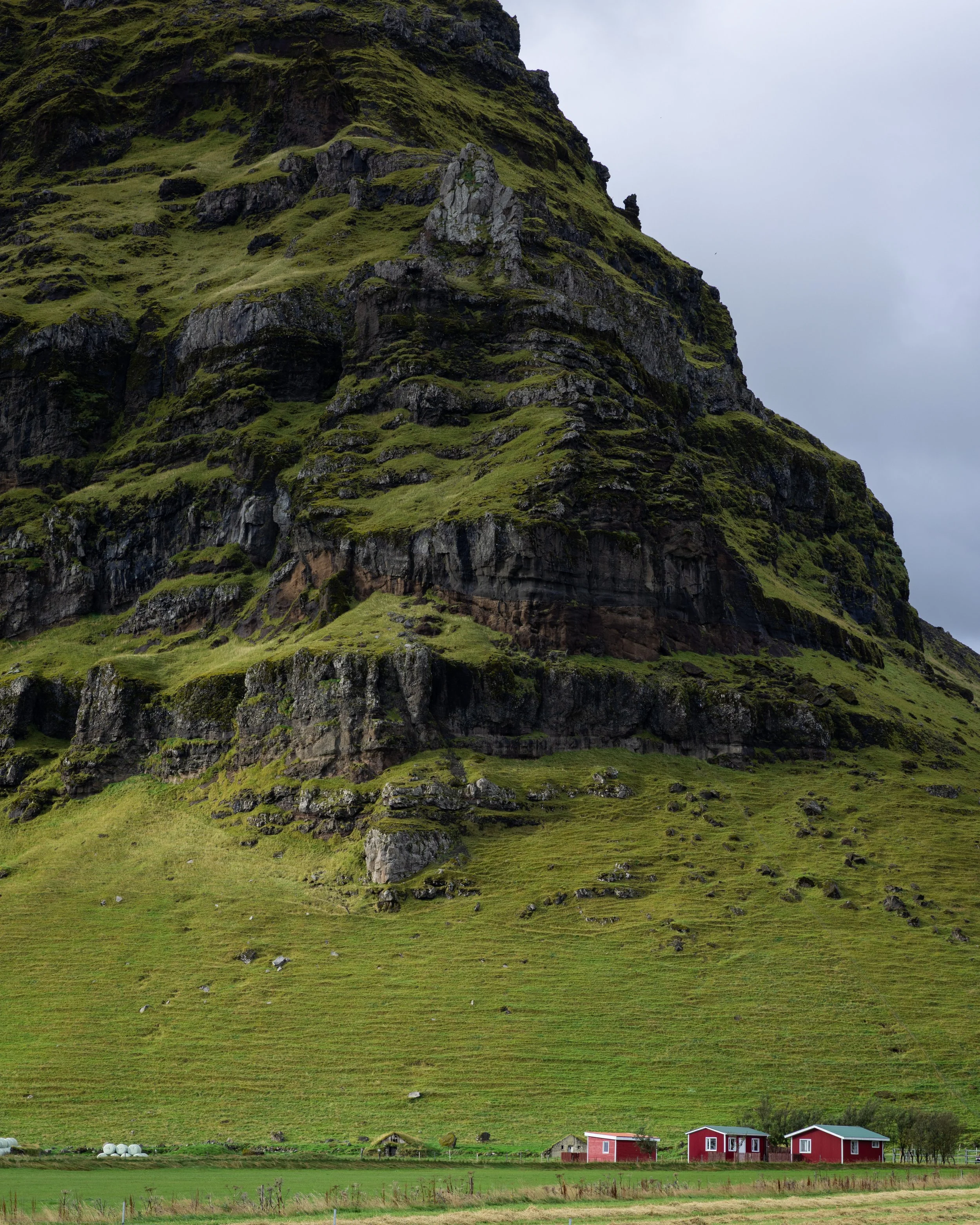 Green hillside with rocky cliff and three red houses at the base under a cloudy sky.
