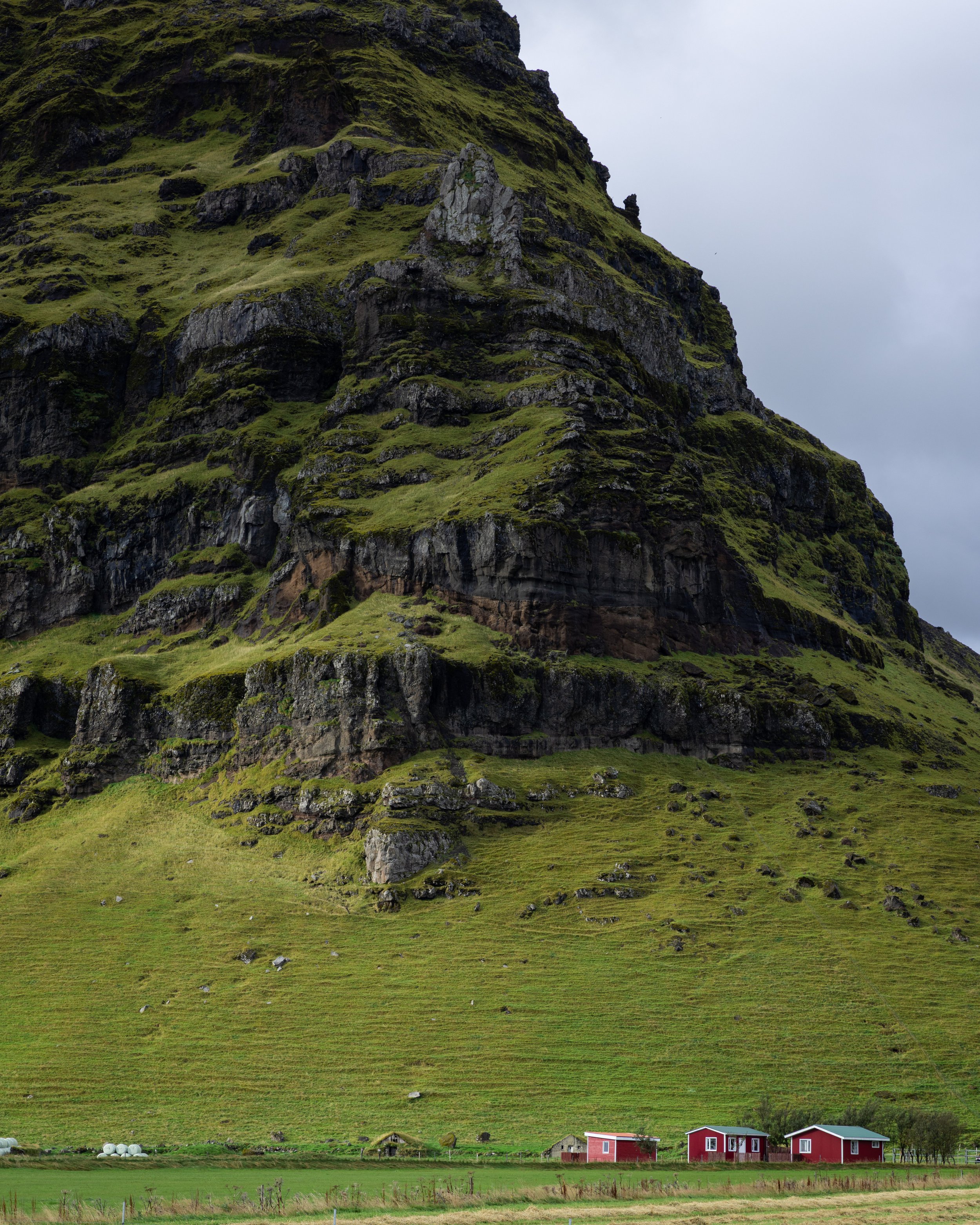 Scenic view of a large green rocky mountain with some small houses at the base, under a cloudy sky.