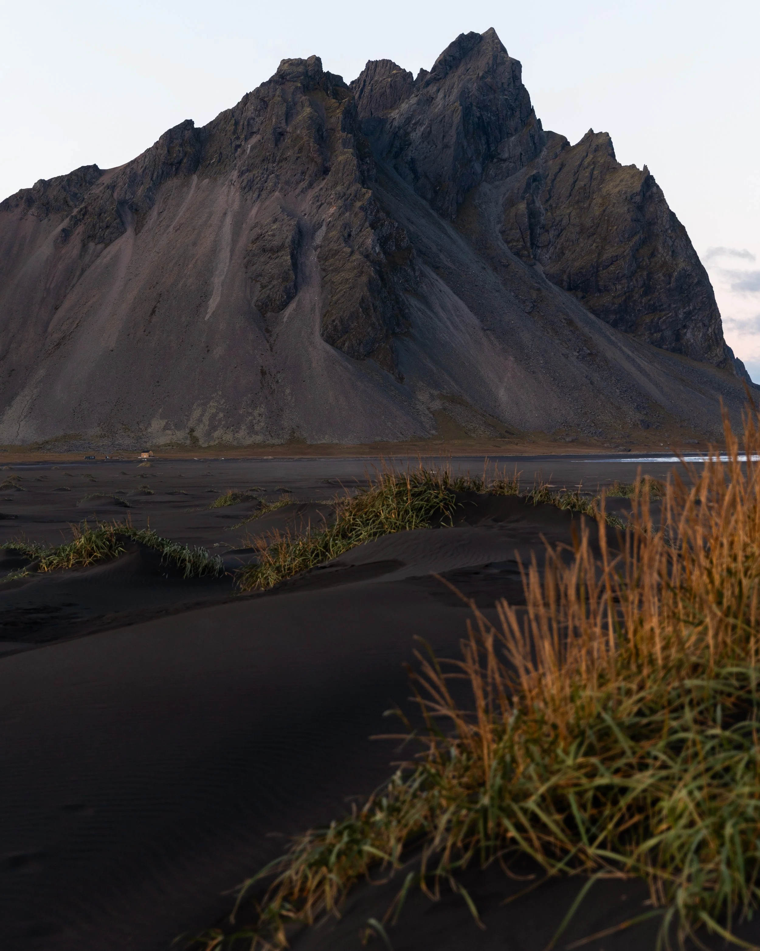 A large mountain with steep rocky slopes and sharp peaks, with dark sand dunes and patches of grass in the foreground.