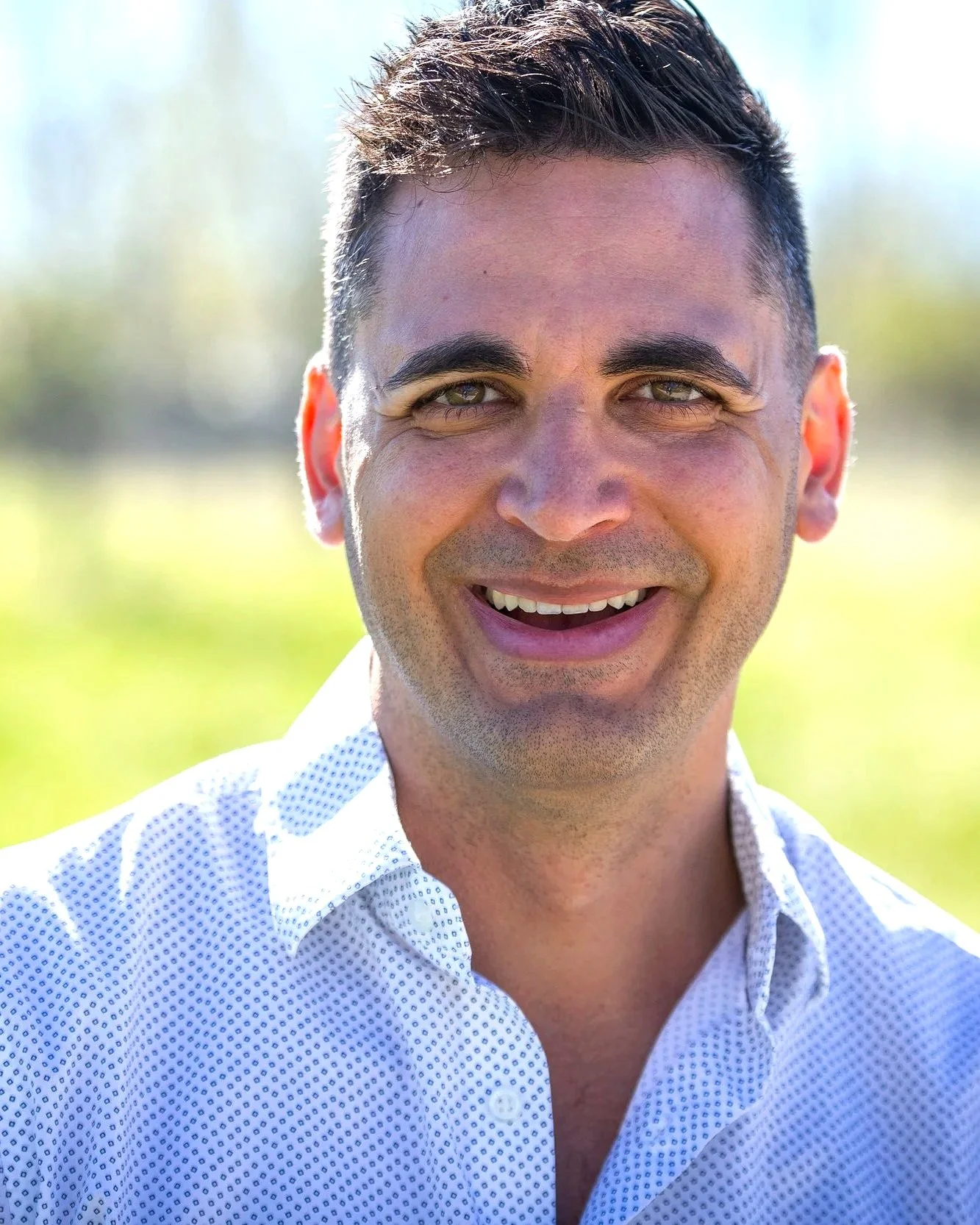 Close-up of a smiling man with short dark hair, light skin, wearing a white shirt with small blue dots, outdoors on a sunny day.