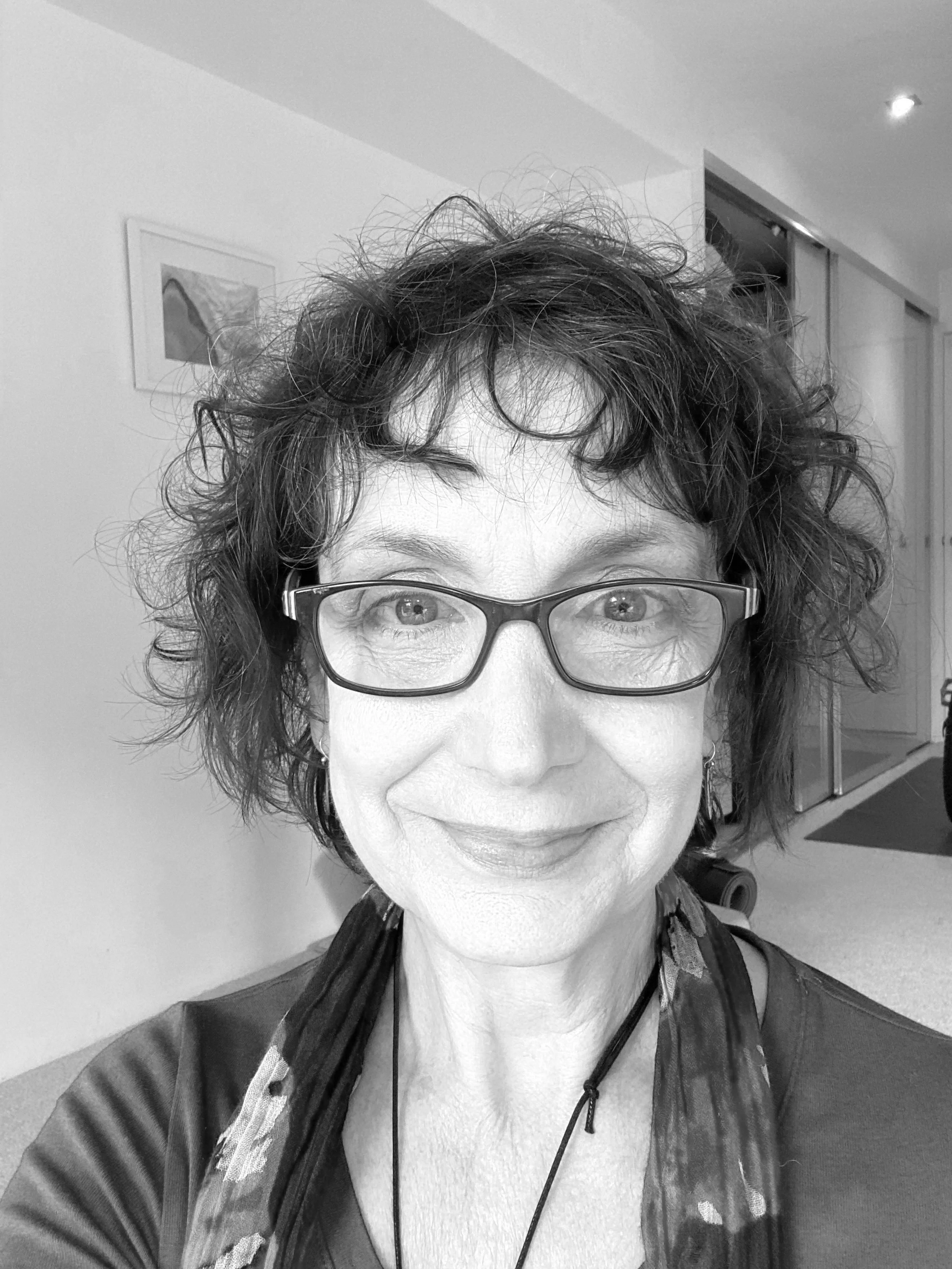 A woman with curly hair and glasses smiling at the camera in an indoor setting.