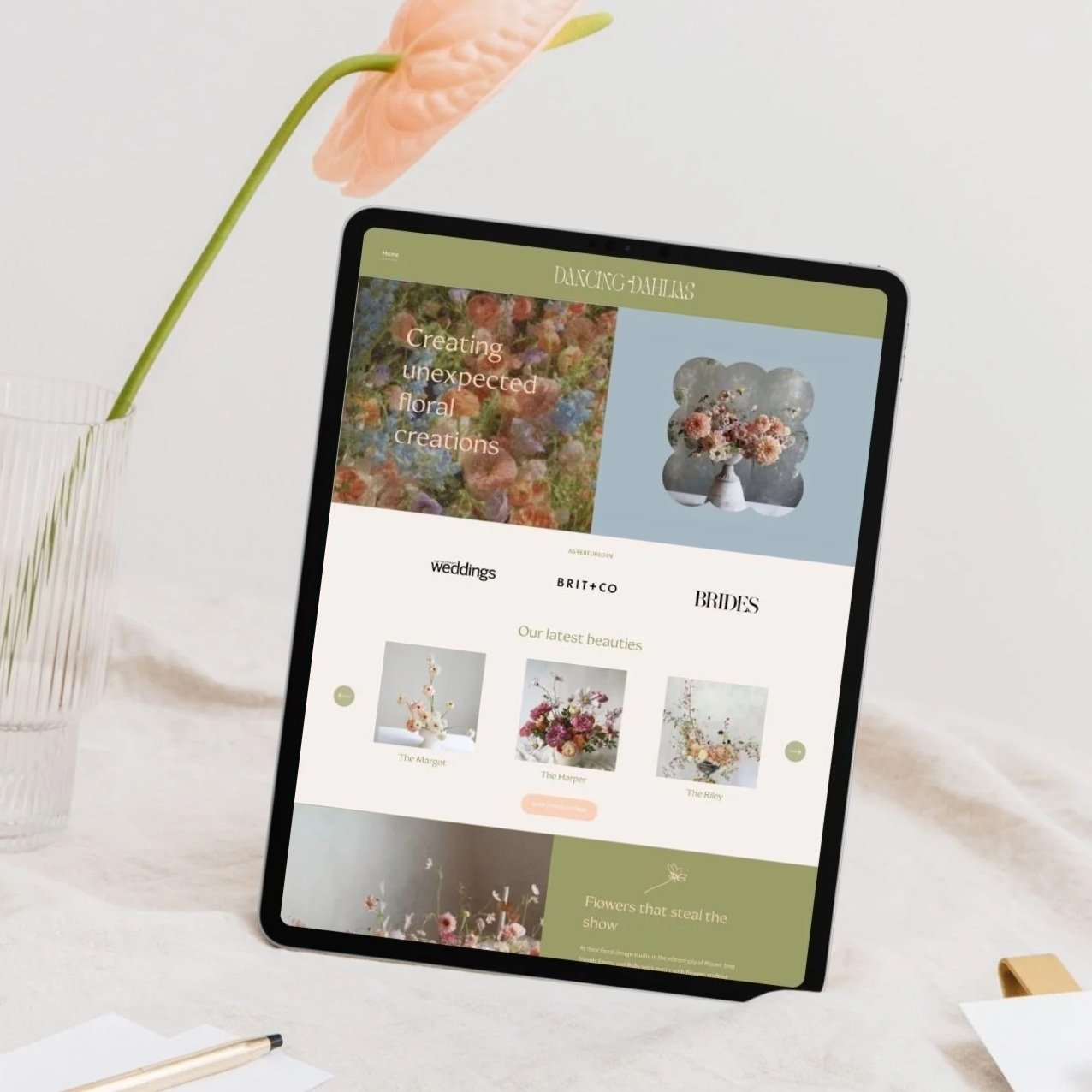 A tablet displaying a floral design website with images of floral arrangements, surrounded by a vase with a peach-colored flower and a gold pen on a white desk.
