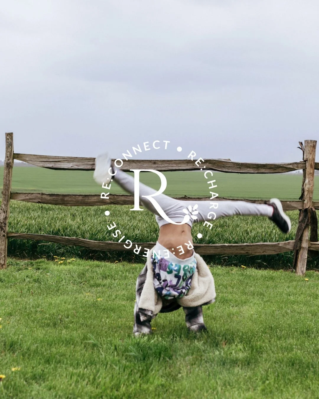 Child doing a handstand on grass in front of a wooden fence, with a cloudy sky and green field in the background.
