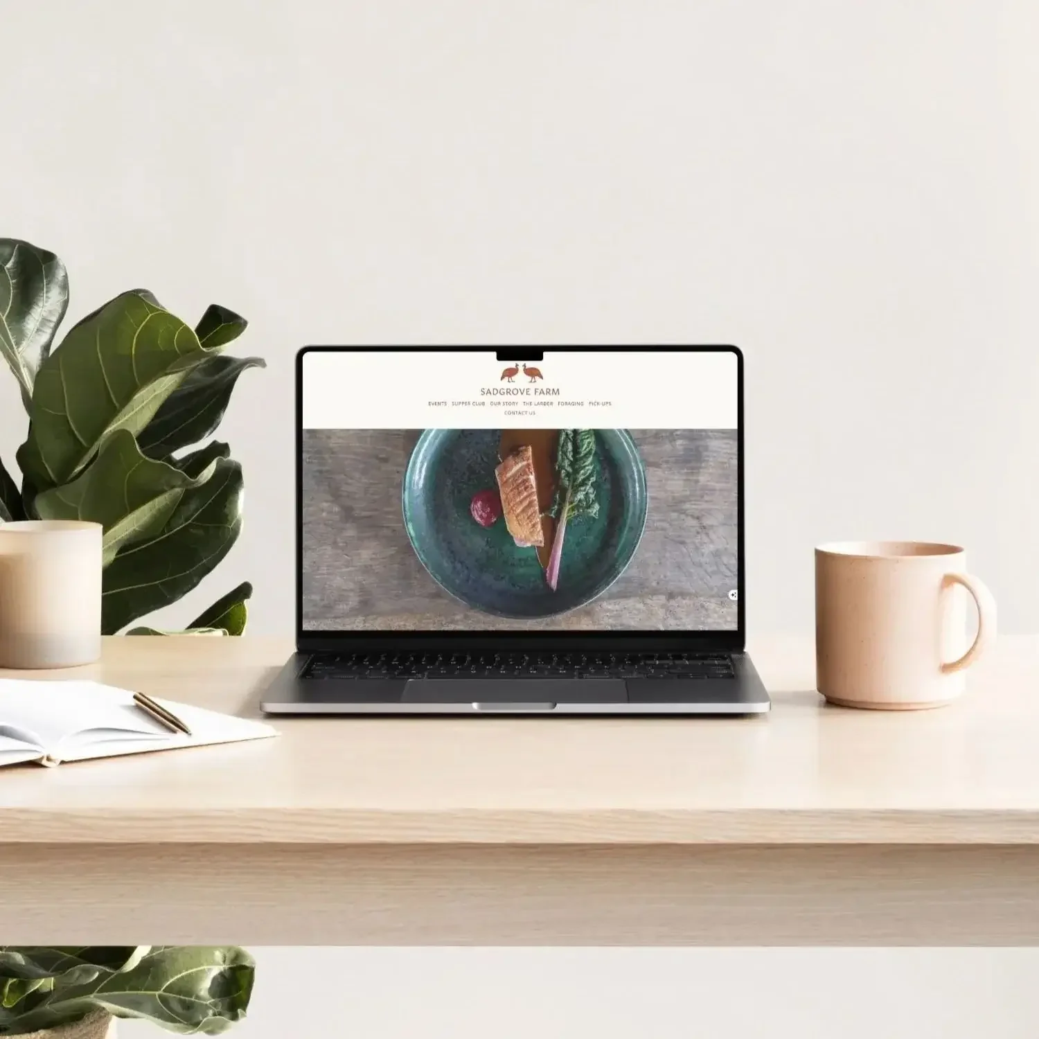 A laptop on a desk displaying the Sadgrove Farm website, with a plant on the left and a pink mug on the right, in a minimalistic setting.