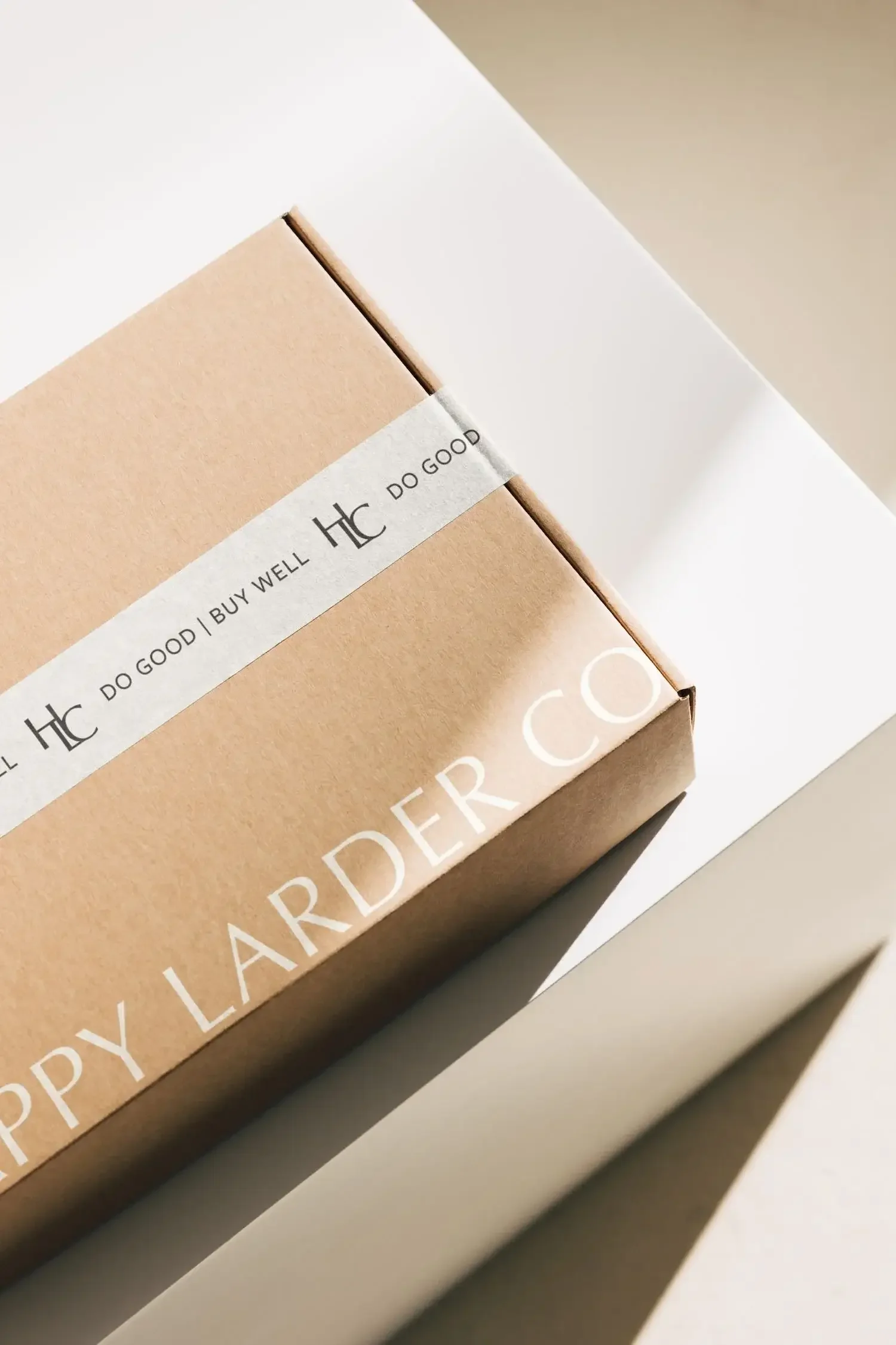 A cardboard box with a white band that says 'DO GOOD | BUY WELL | DO GOOD' resting on a white surface, with a shadow cast across it.