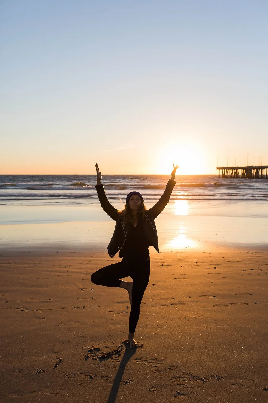 Person practicing yoga on the beach during sunset, standing on one leg with arms raised, near ocean waves and a pier in the background.
