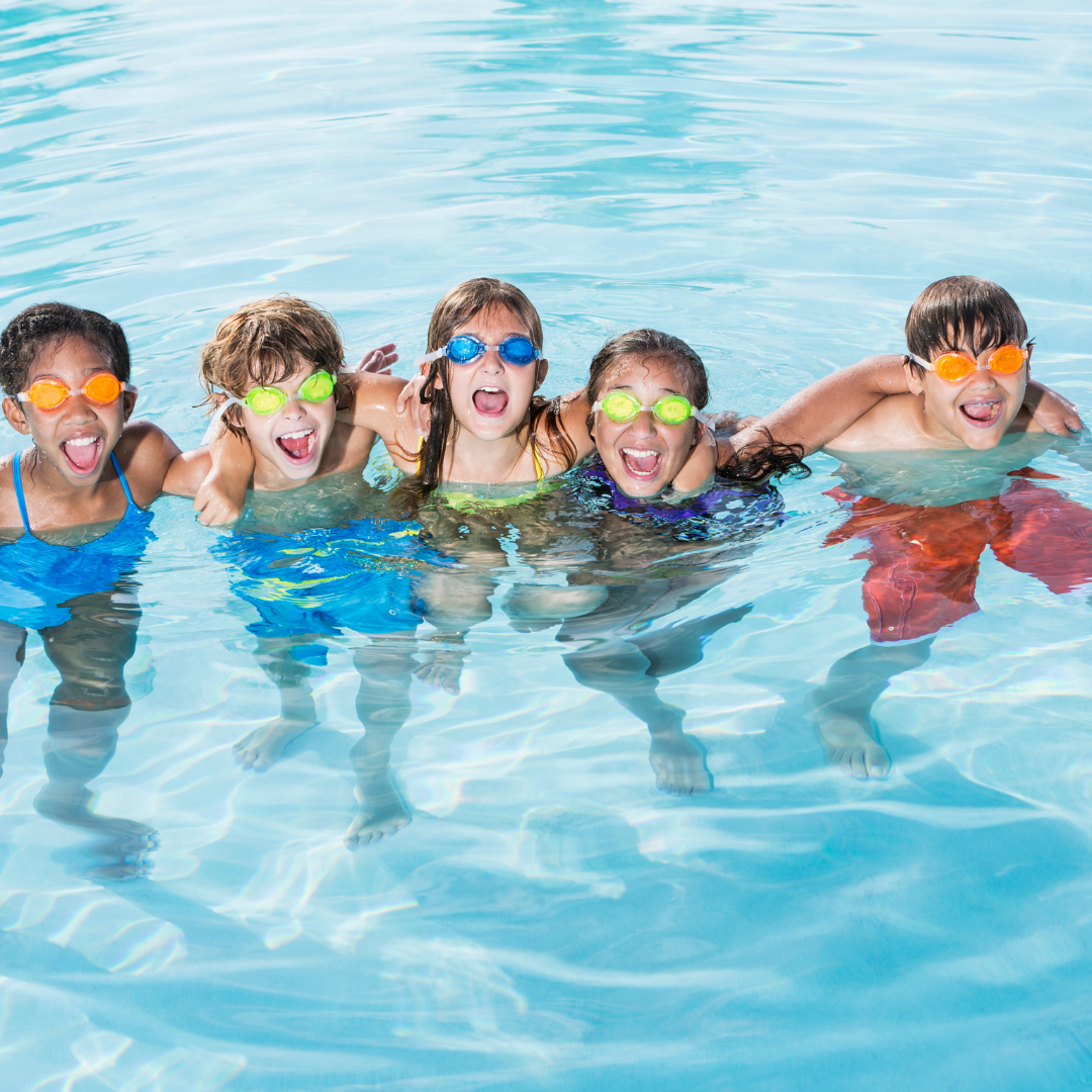 Group of five children wearing colorful goggles and swimsuits, smiling and playing in a swimming pool.