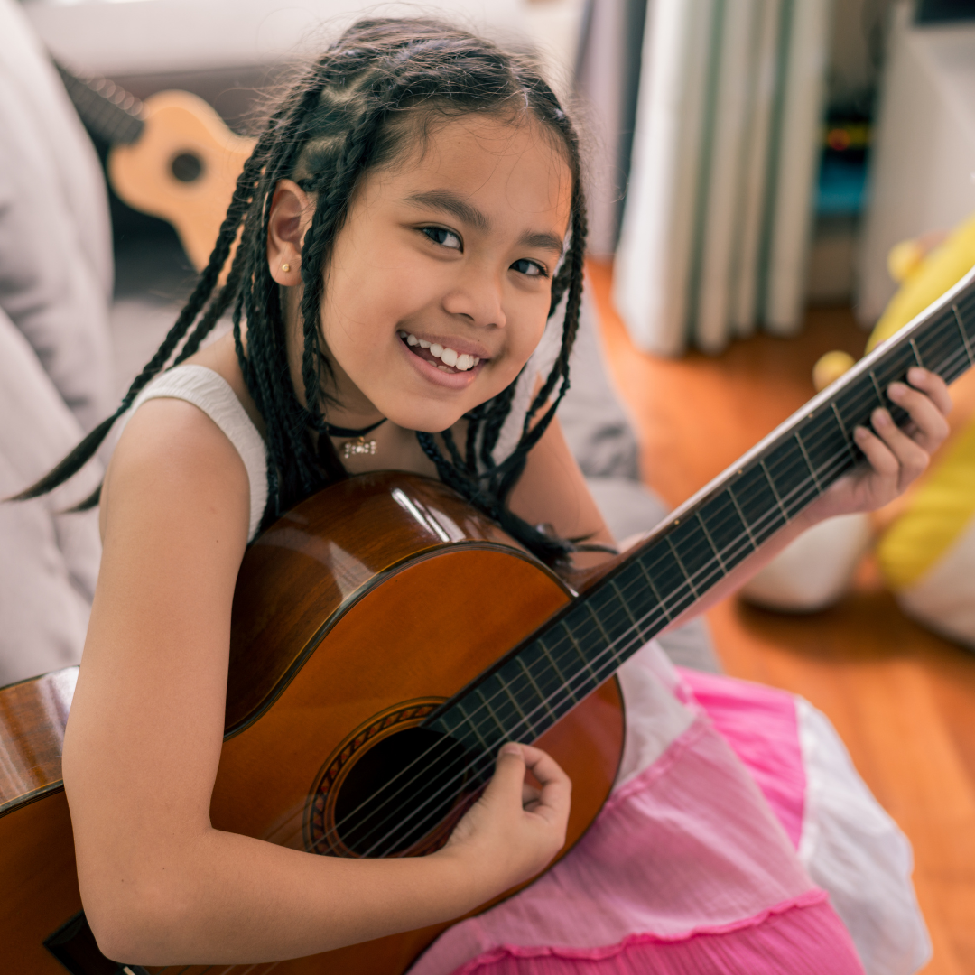 A young girl with braided hair smiling while holding a guitar indoors.