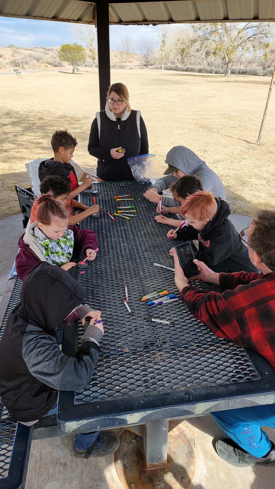 A group of children and adults sitting at a picnic table outdoors under a pavilion, engaging in drawing or coloring activities with markers and pens. One adult is taking a photo with a smartphone, while another stands at the end of the table watching