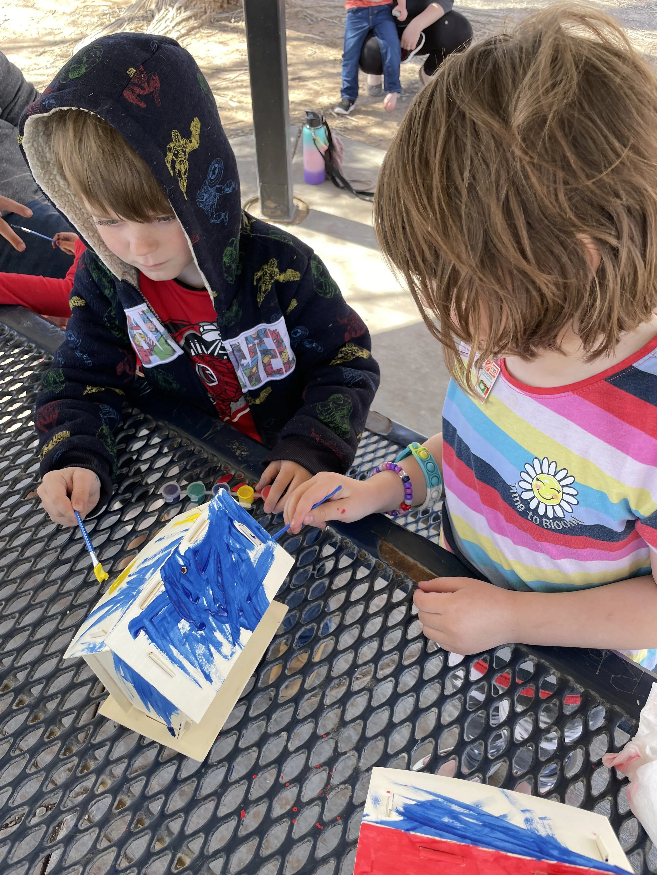Two children painting a small object with blue paint at a metal picnic table outdoors.