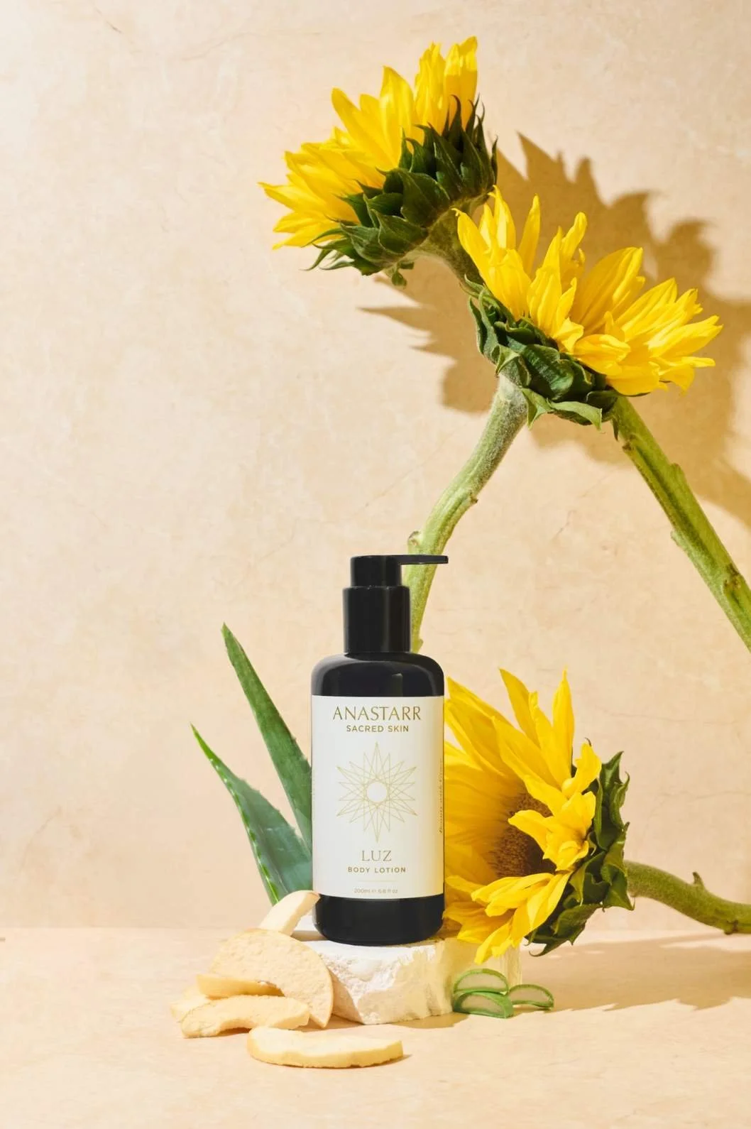 Skincare product photography of a body lotion bottle surrounded by pieces of aloe and apple slices along with sunflowers in front of a beige stone background with warm sunny lighting
